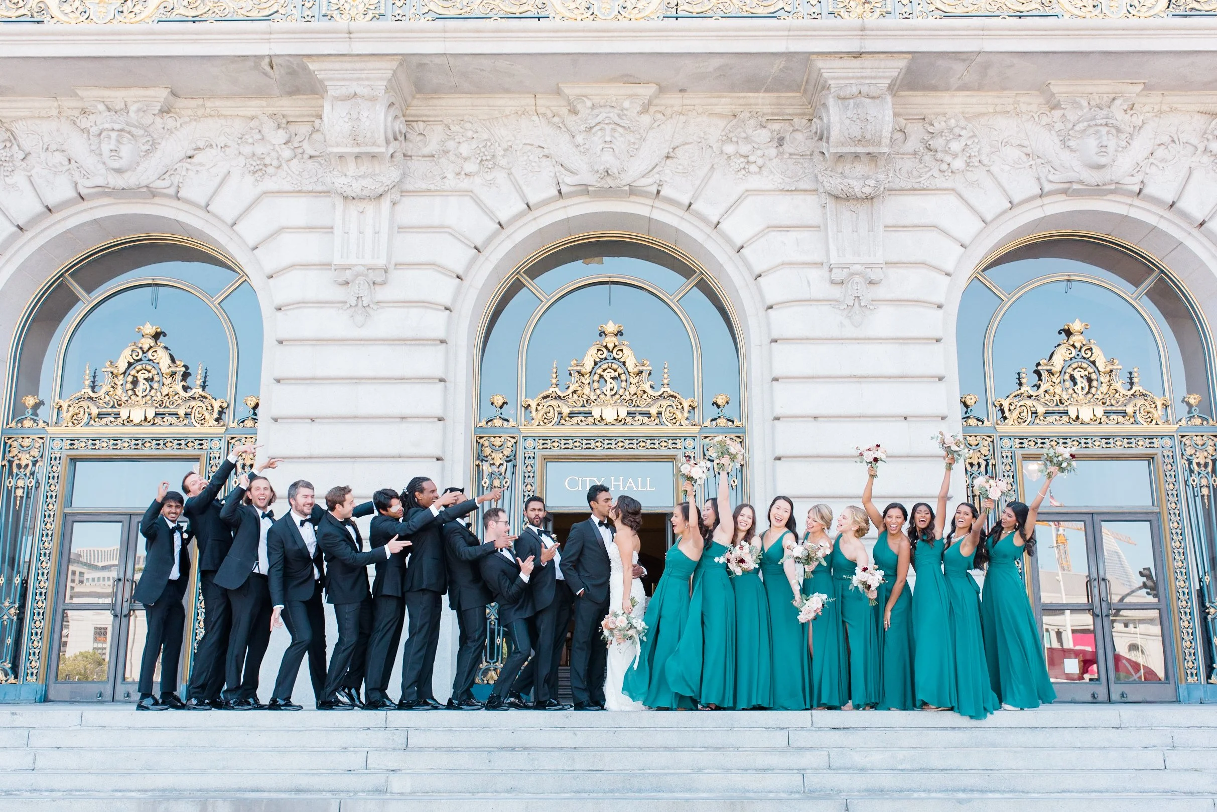 Full bridal party celebrating on the front steps of San Francisco City Hall with the couple kissing in the center.