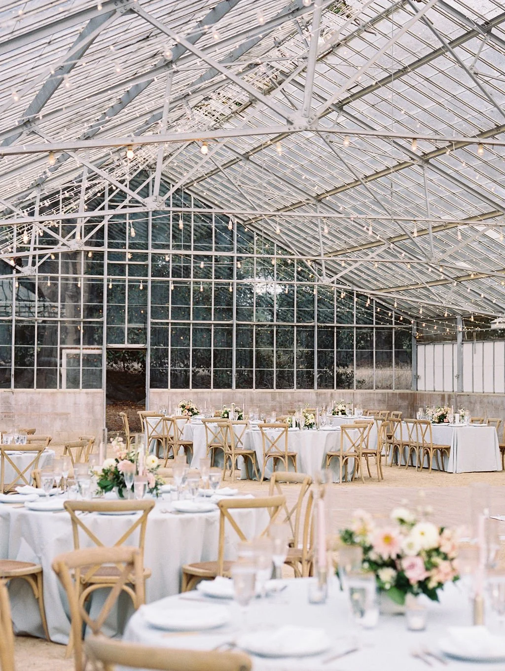 Elegant wedding reception inside the Dos Pueblos Orchid Farm greenhouse with round tables, wooden chairs, and soft café lighting in Santa Barbara.