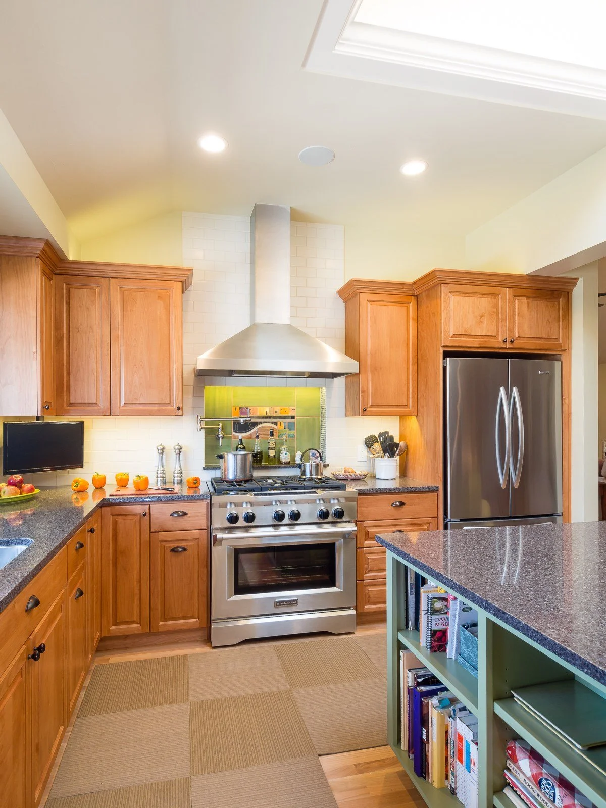 Natural wood kitchen cabinets around a stainless range and hood, with a green tile backsplash and stainless refrigerator