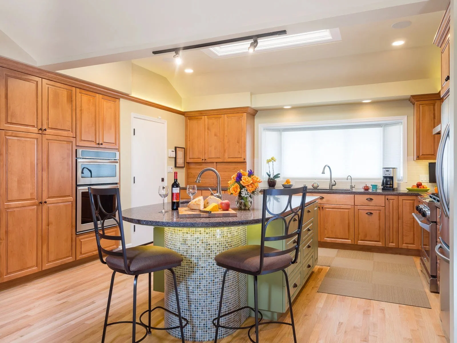 Wood kitchen cabinets with a curved island wrapped in green mosaic tile, bar stools, and double wall ovens