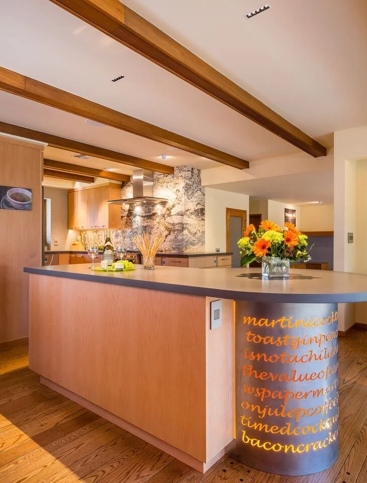 Curved light wood kitchen island under exposed beams, with a stone backsplash and range hood behind