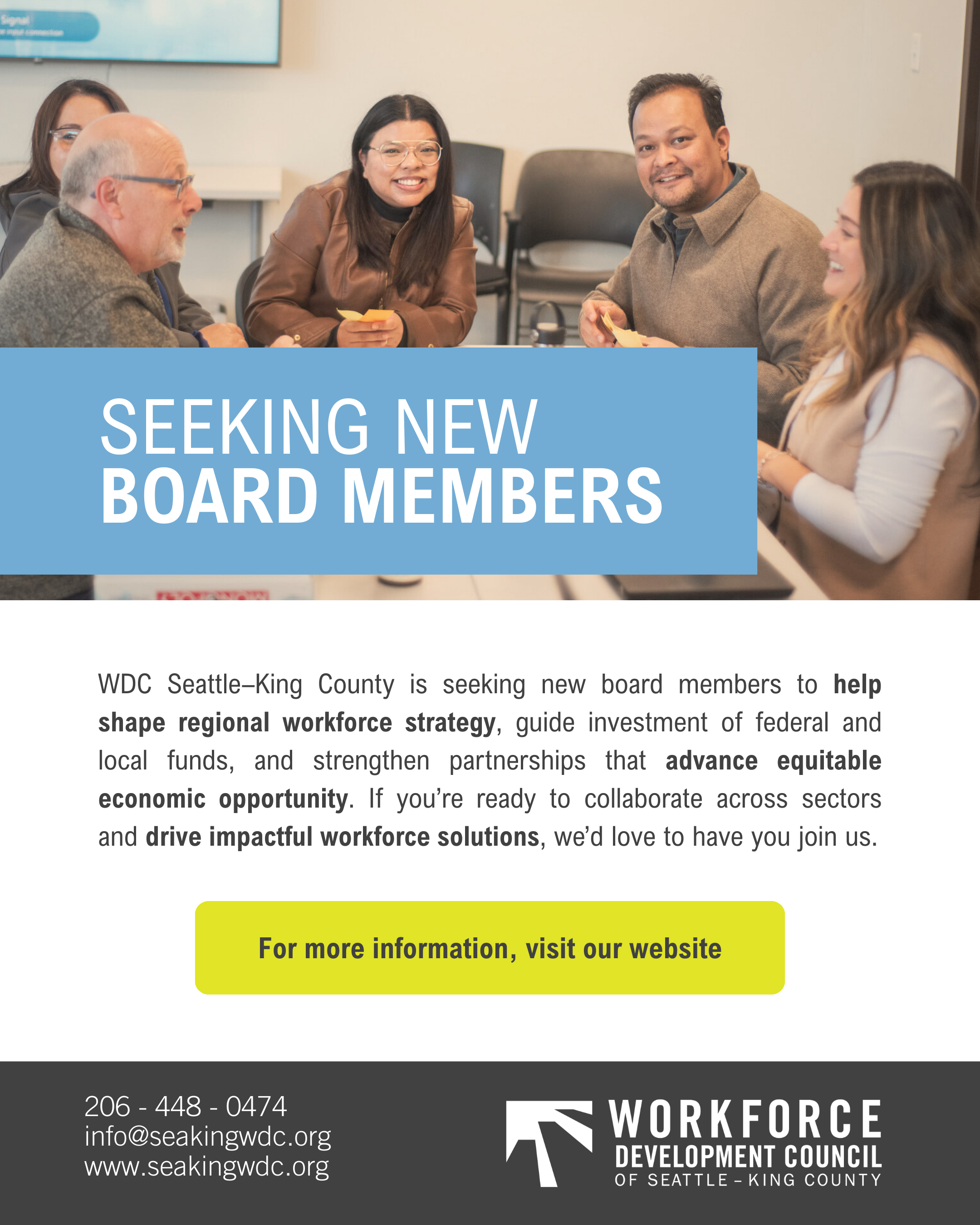 Group of five diverse people sitting around a table in a meeting room, smiling and talking. A large screen is visible in the background.