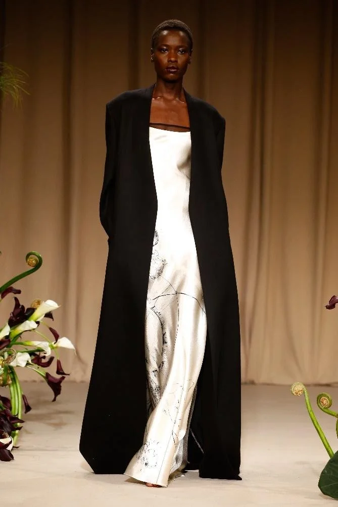Model wearing Jason Wu Fall Winter 2023 outfit consisting of a black coat over a white silk dress featuring a sketch design