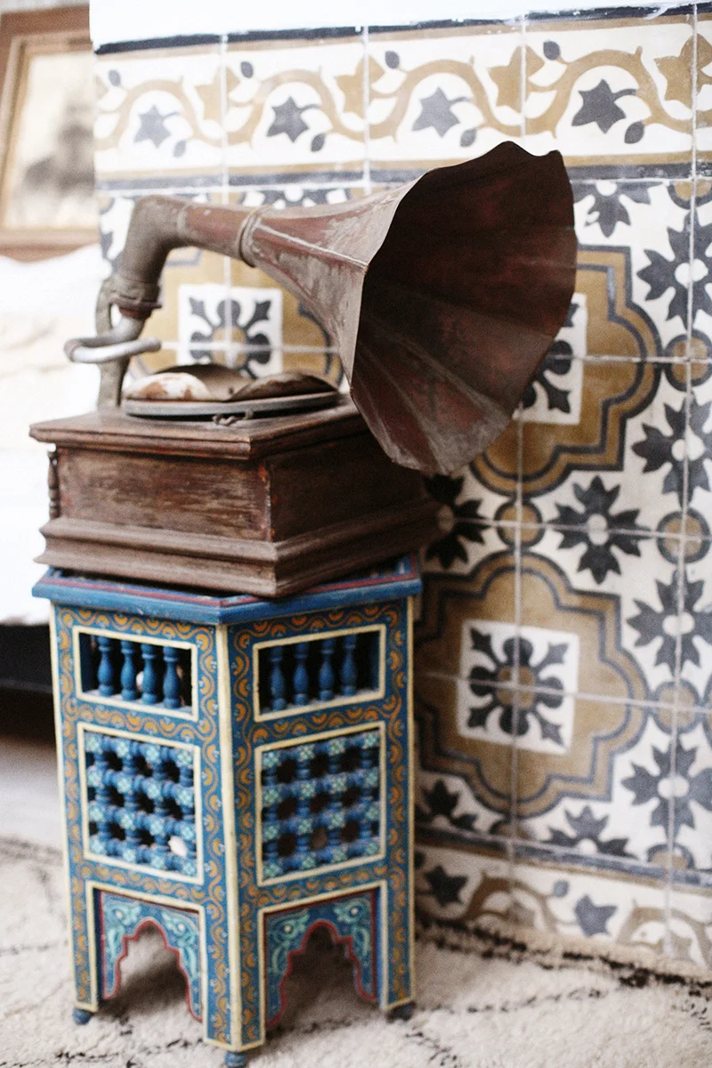 Vintage gramophone with a dark wooden finish, placed on a decorative blue and gold stand, against a background of patterned tile wall.
