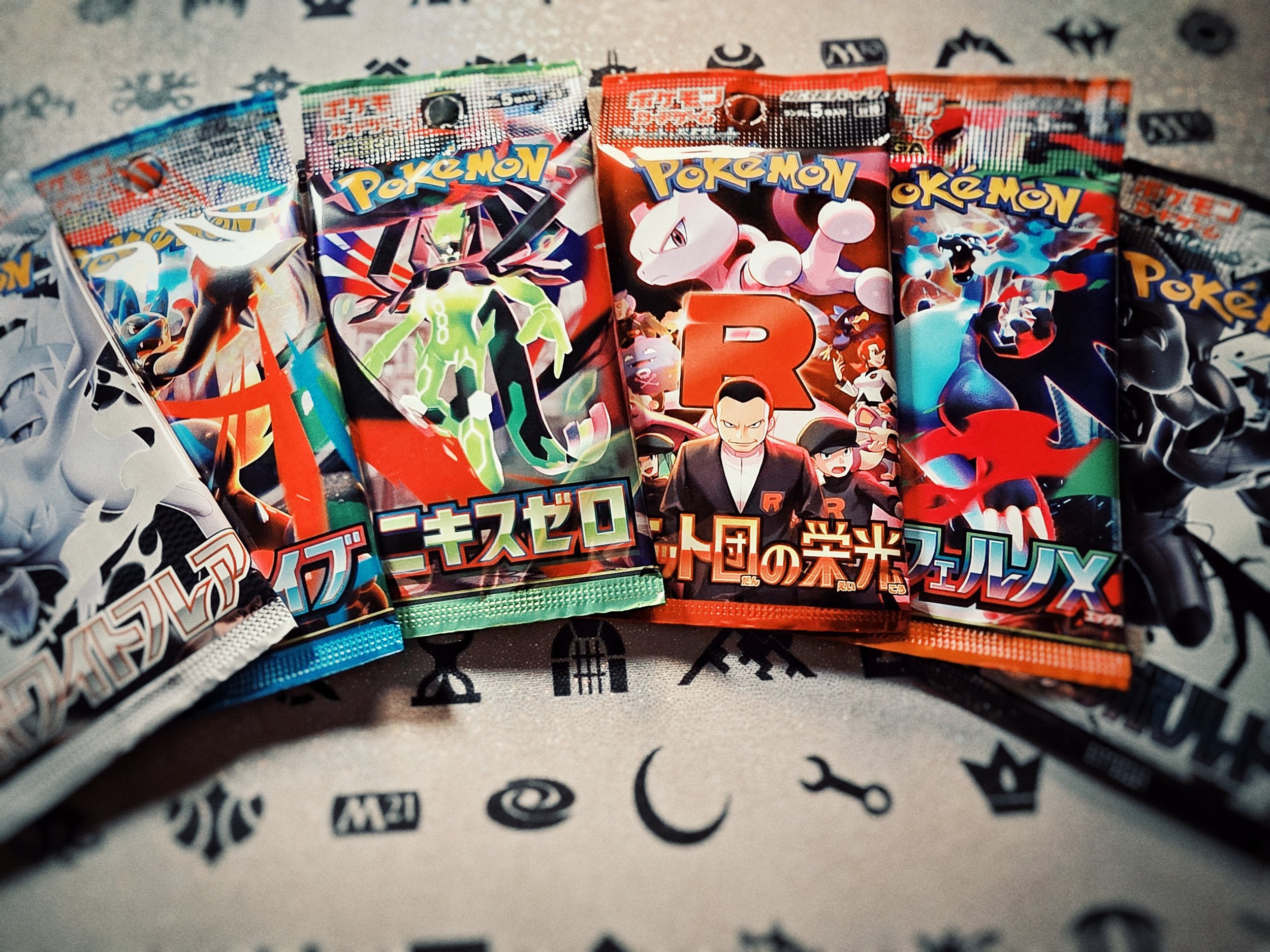 Cinque bustine di carte da gioco Pokémon disposte su un tessuto con simboli e scritte nere.