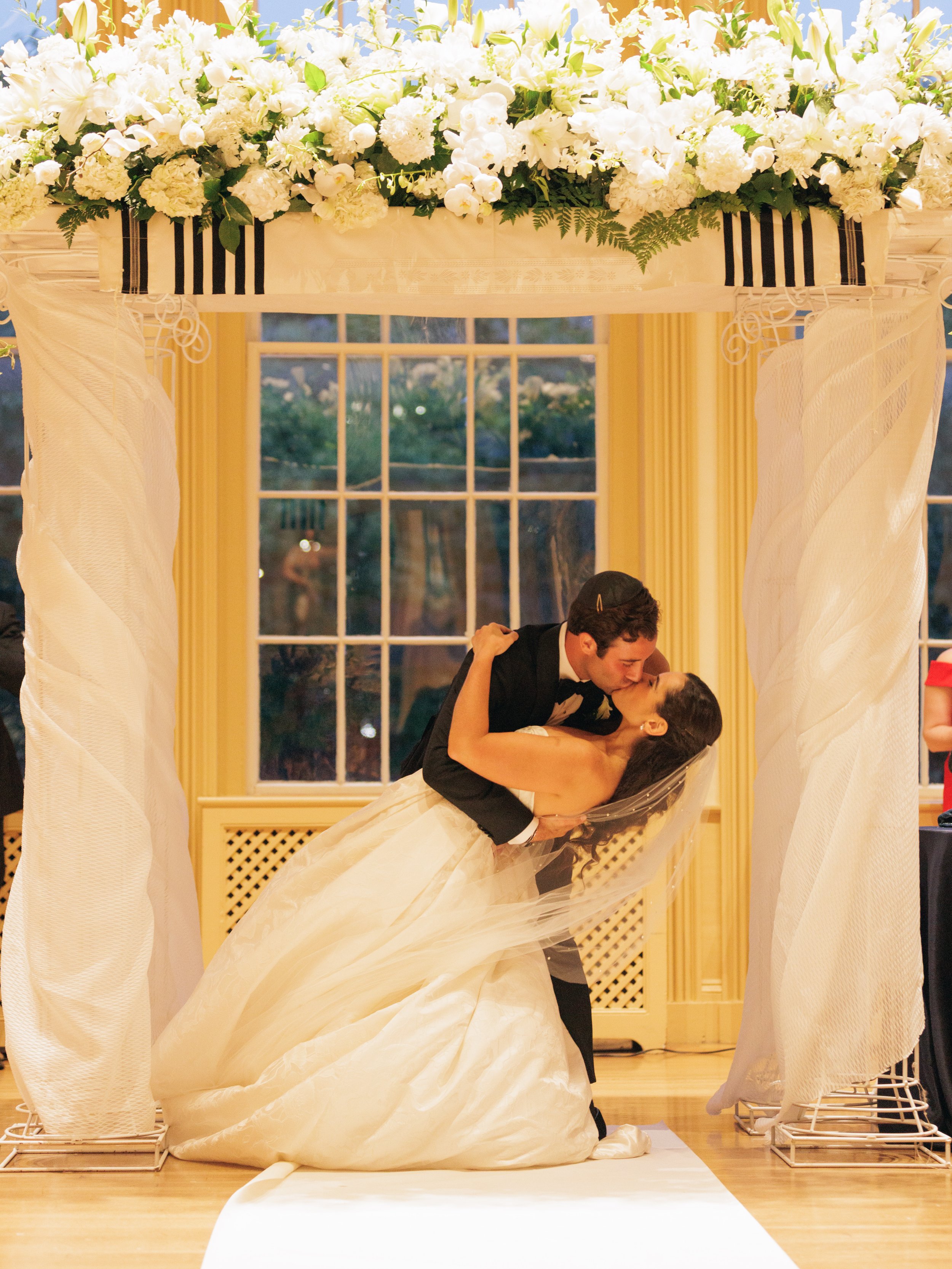 A bride and groom sharing a kiss under a floral wedding arch with white flowers and greenery in a room with yellow walls and a window in the background.