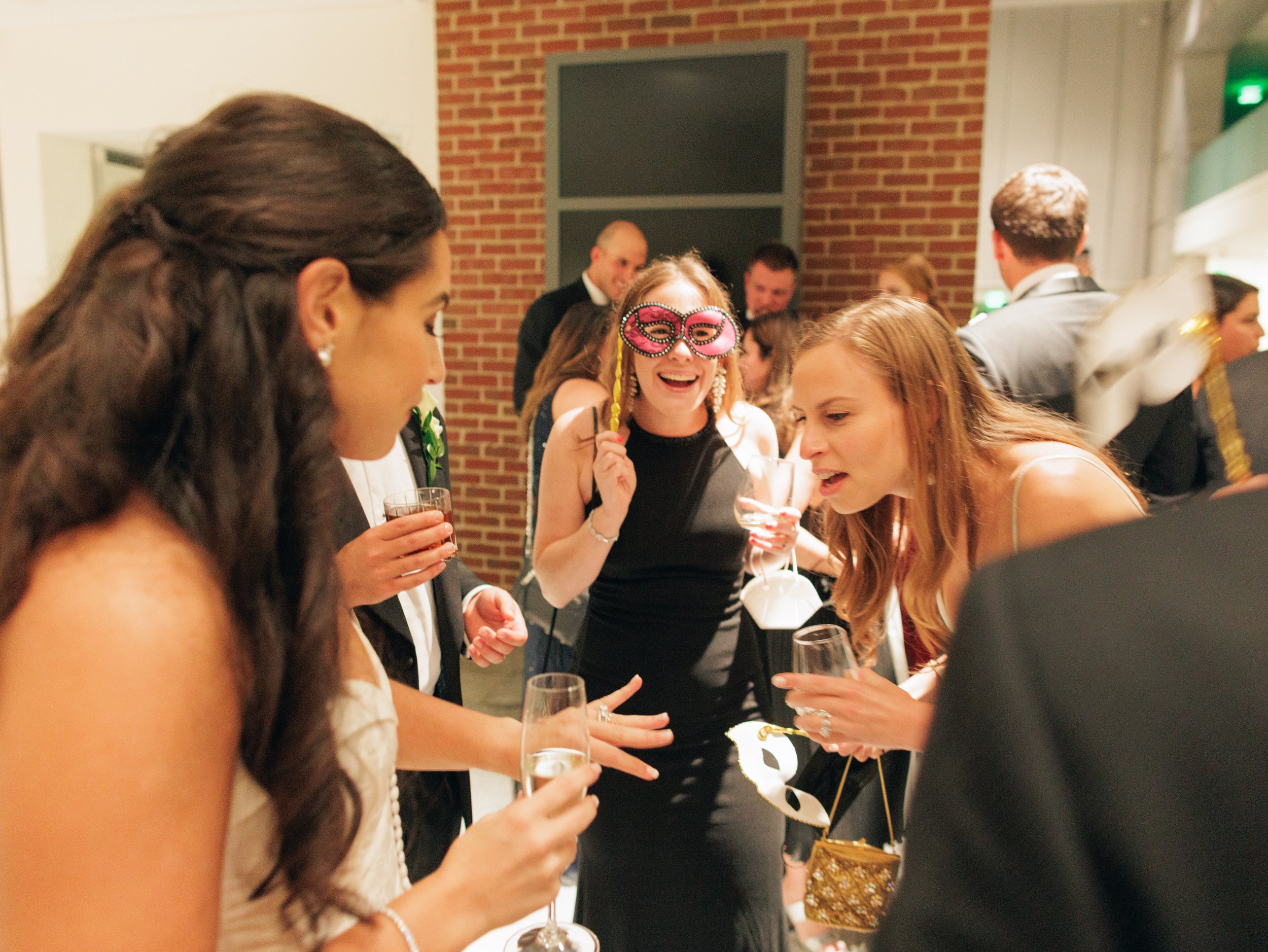 People dressed in formal attire at a party, with one woman wearing a pink masquerade mask, holding drinks, and smiling in a social setting with a brick wall in the background.