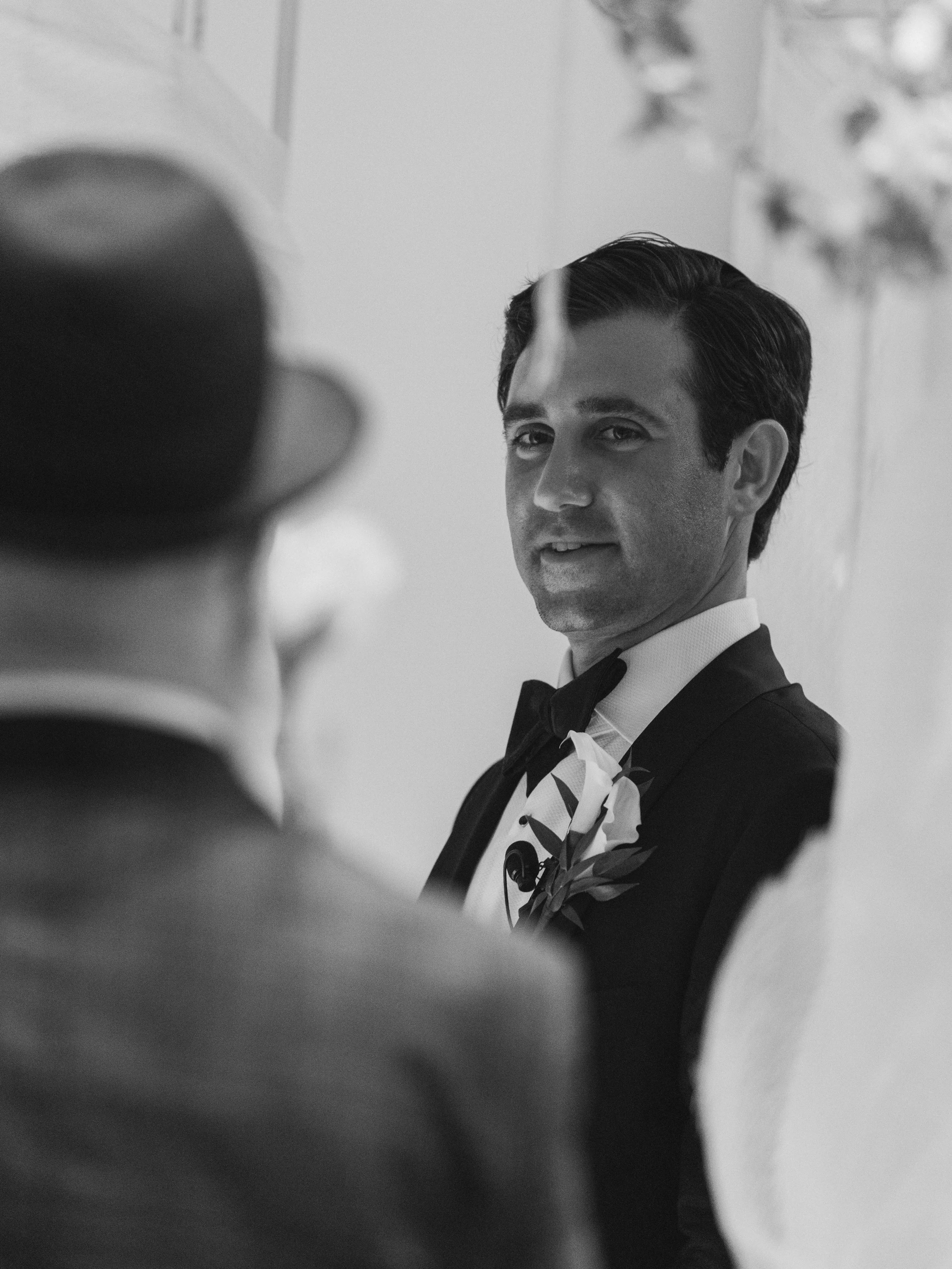 A black-and-white photo of a man in a tuxedo with a bow tie, looking into the camera with a slight smile, and a boutonniere on his left lapel.
