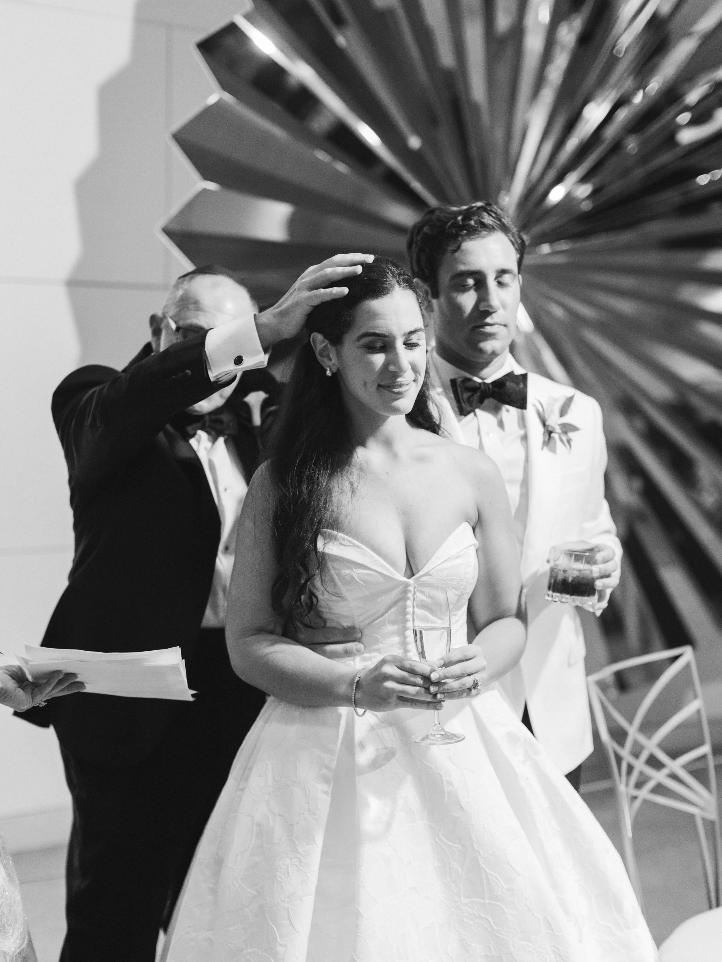 Black and white photo of a newlywed couple at a wedding reception, with a man in a tuxedo touching the bride's head, the bride holding a glass of champagne, and another man in a tuxedo beside her, all with a decorative backdrop.