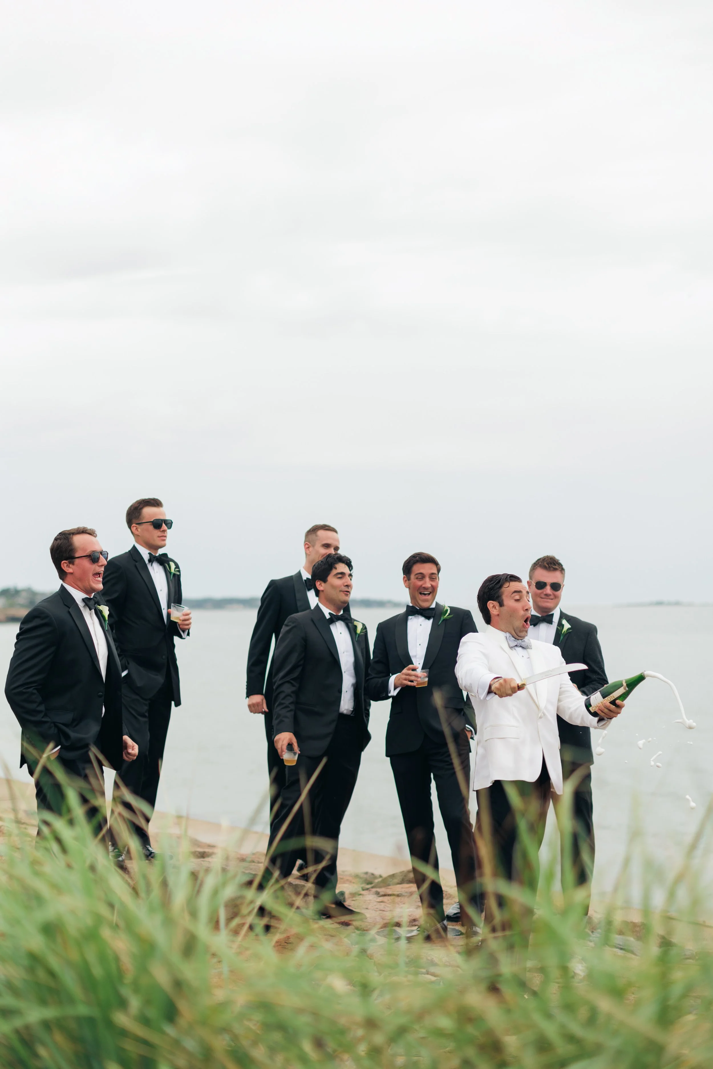 A group of men dressed in tuxedos with sunglasses walking on the beach, with some holding drinks and one man in a white tuxedo opening a bottle of champagne.