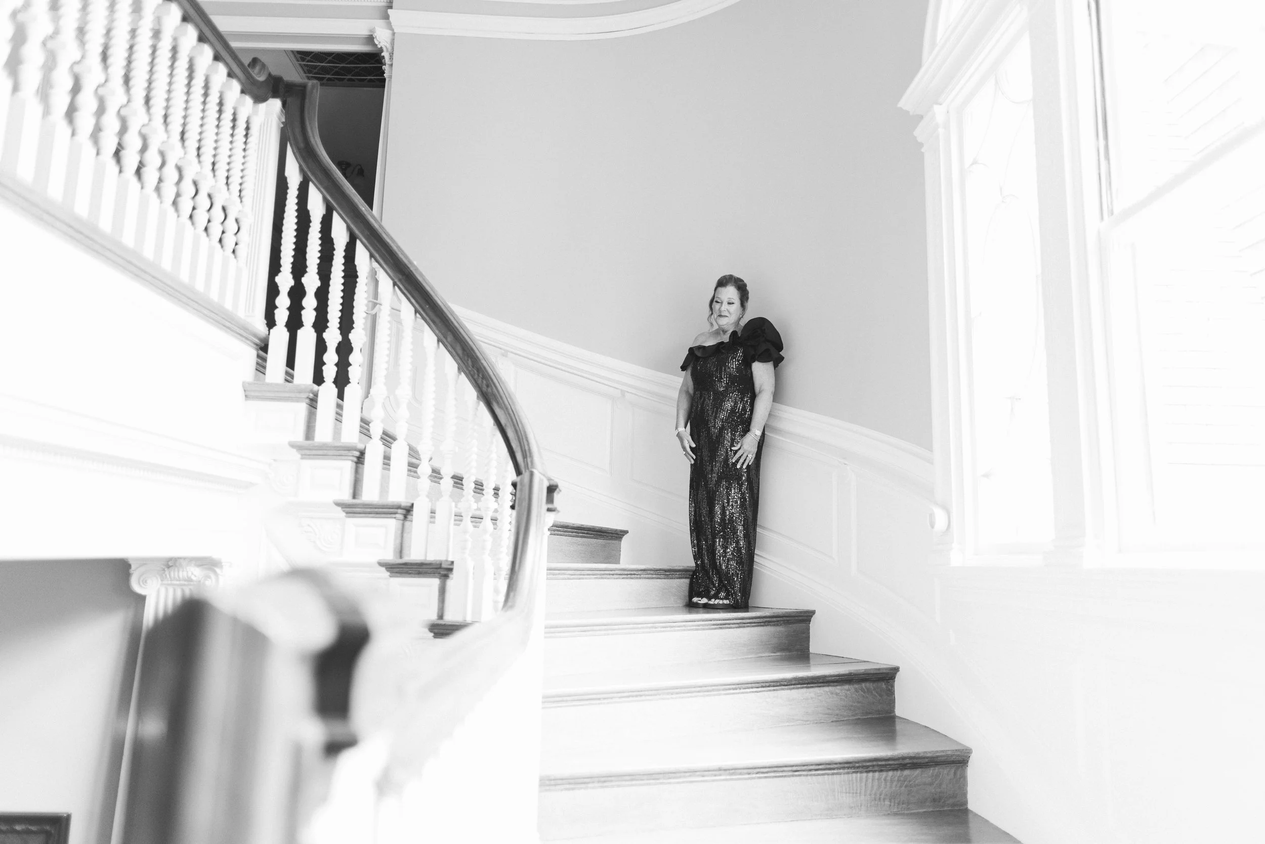 A woman in a glamorous, sparkling dress standing on a staircase in a well-lit, elegant interior with detailed wall moldings.