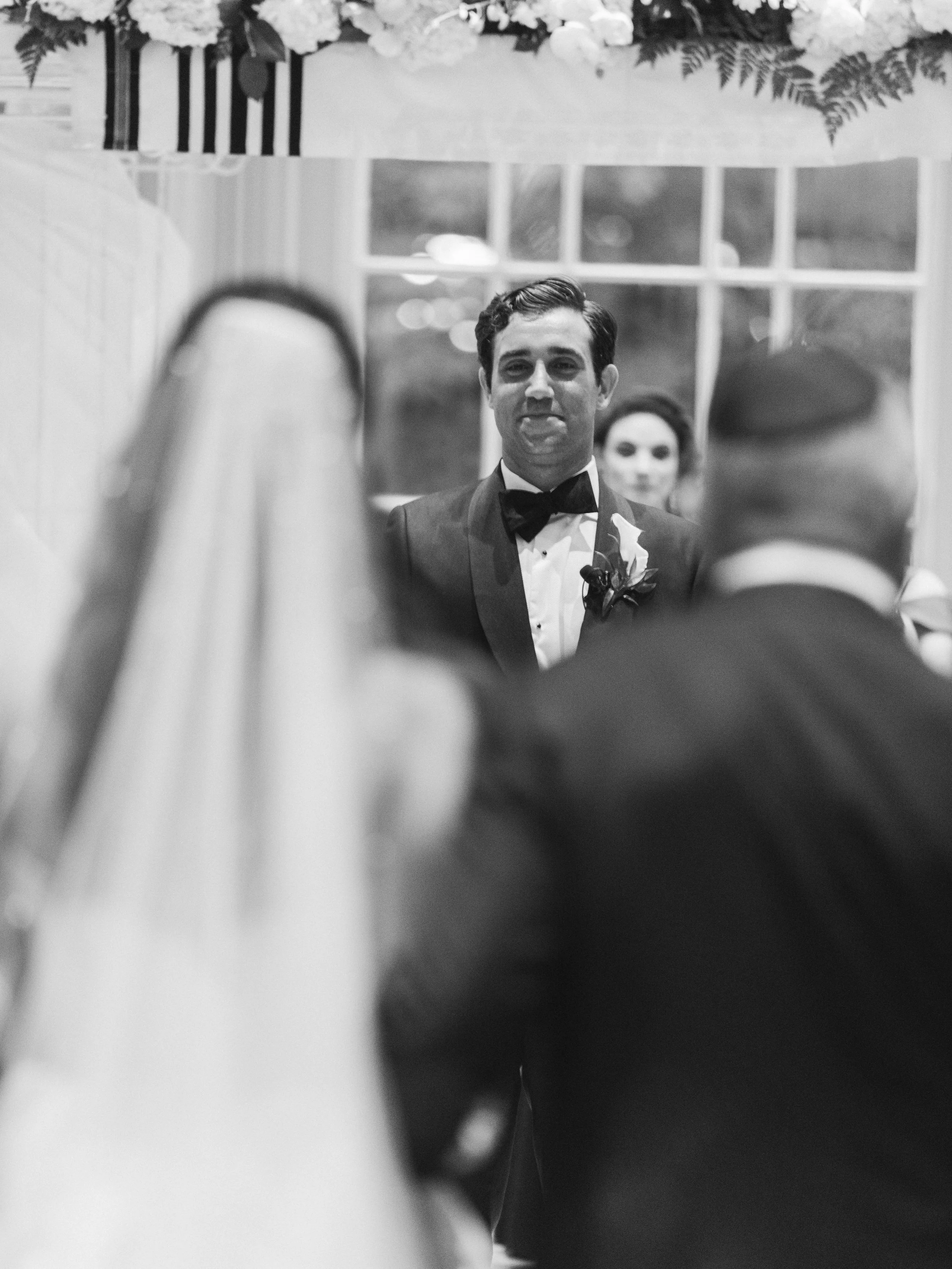 Black and white photo of a man in a tuxedo with a bow tie, smiling, with a woman behind him, and floral decorations above, possibly at a formal event or wedding.