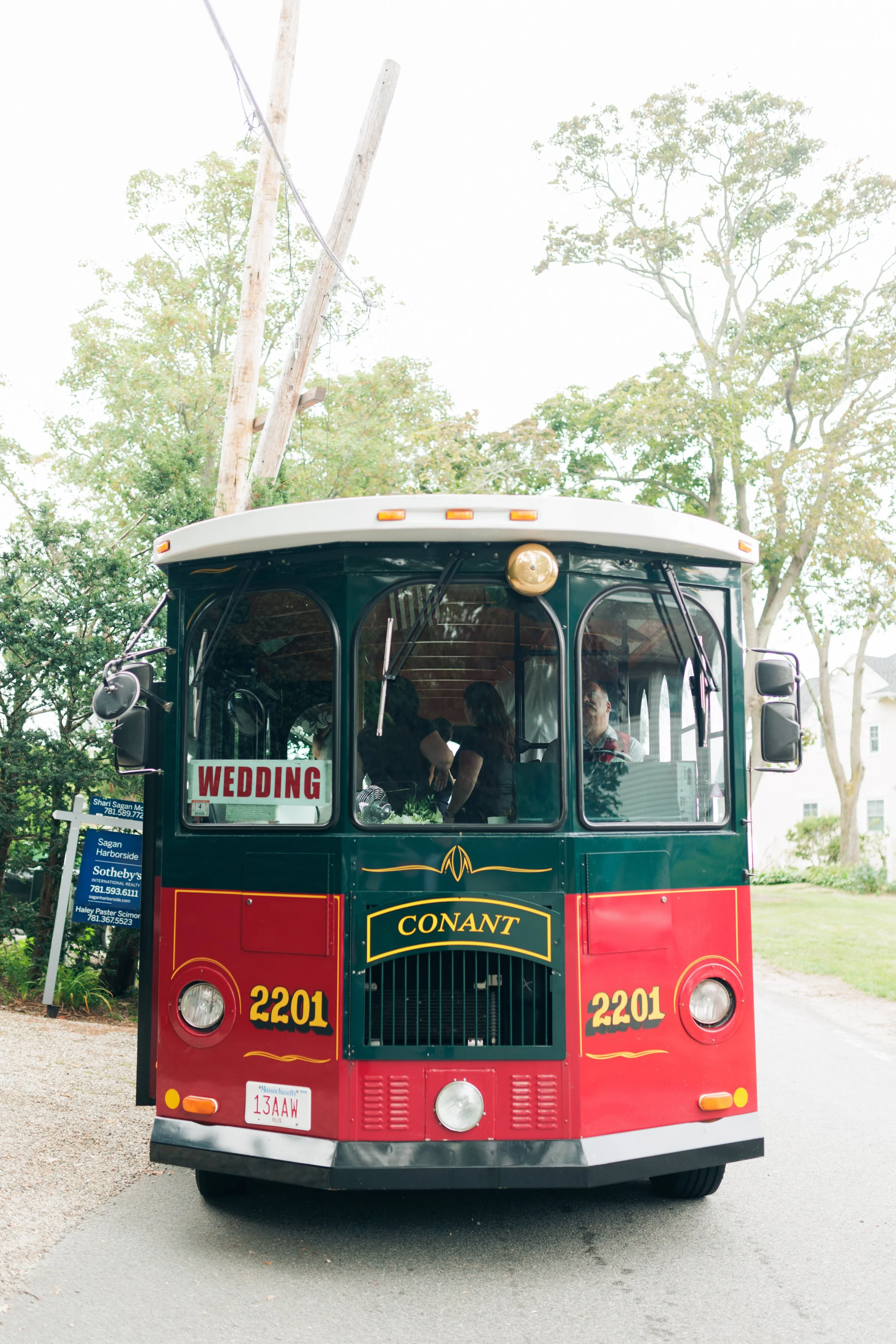 Red and black trolley bus with a sign that says 'Wedding' on the front, parking on a street with trees and a sign for real estate agency in the background.