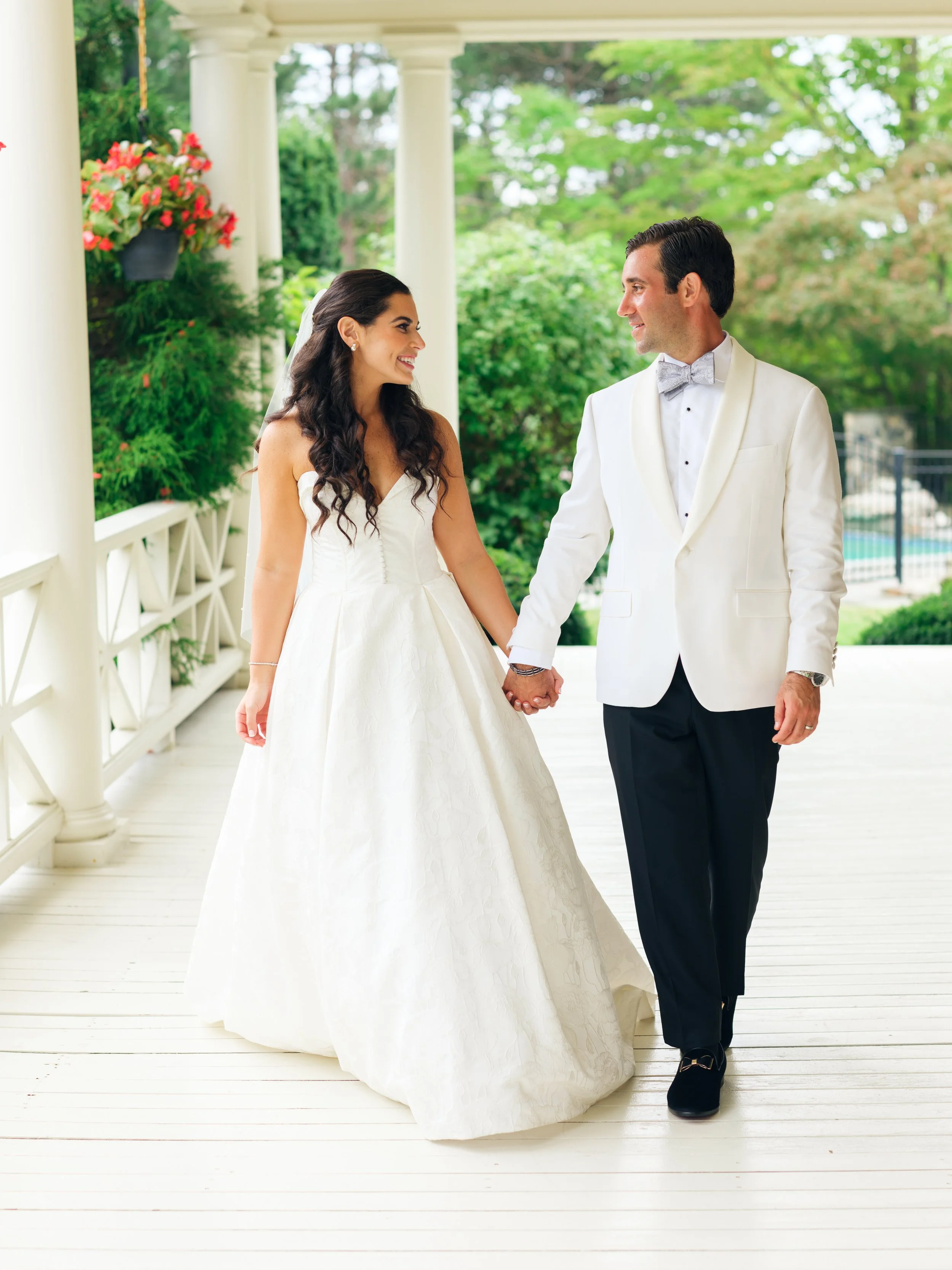 A bride and groom holding hands and smiling at each other on a white porch with green trees in the background.