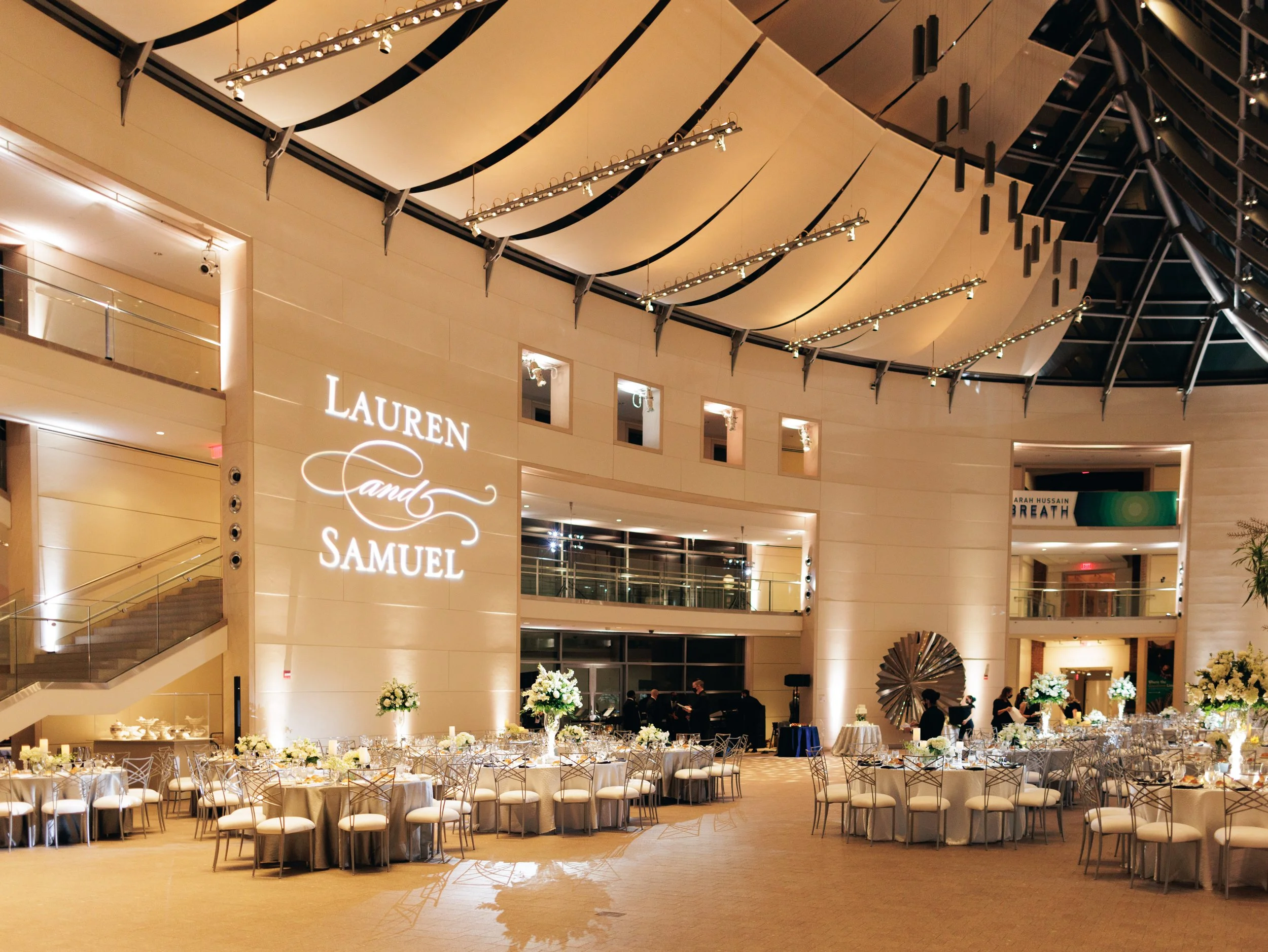 An elegant event space decorated for a wedding reception, featuring round tables with white and silver tablecloths, floral centerpieces, and modern white chairs. The upper part of the room has a high, curved ceiling with black accents and suspended l