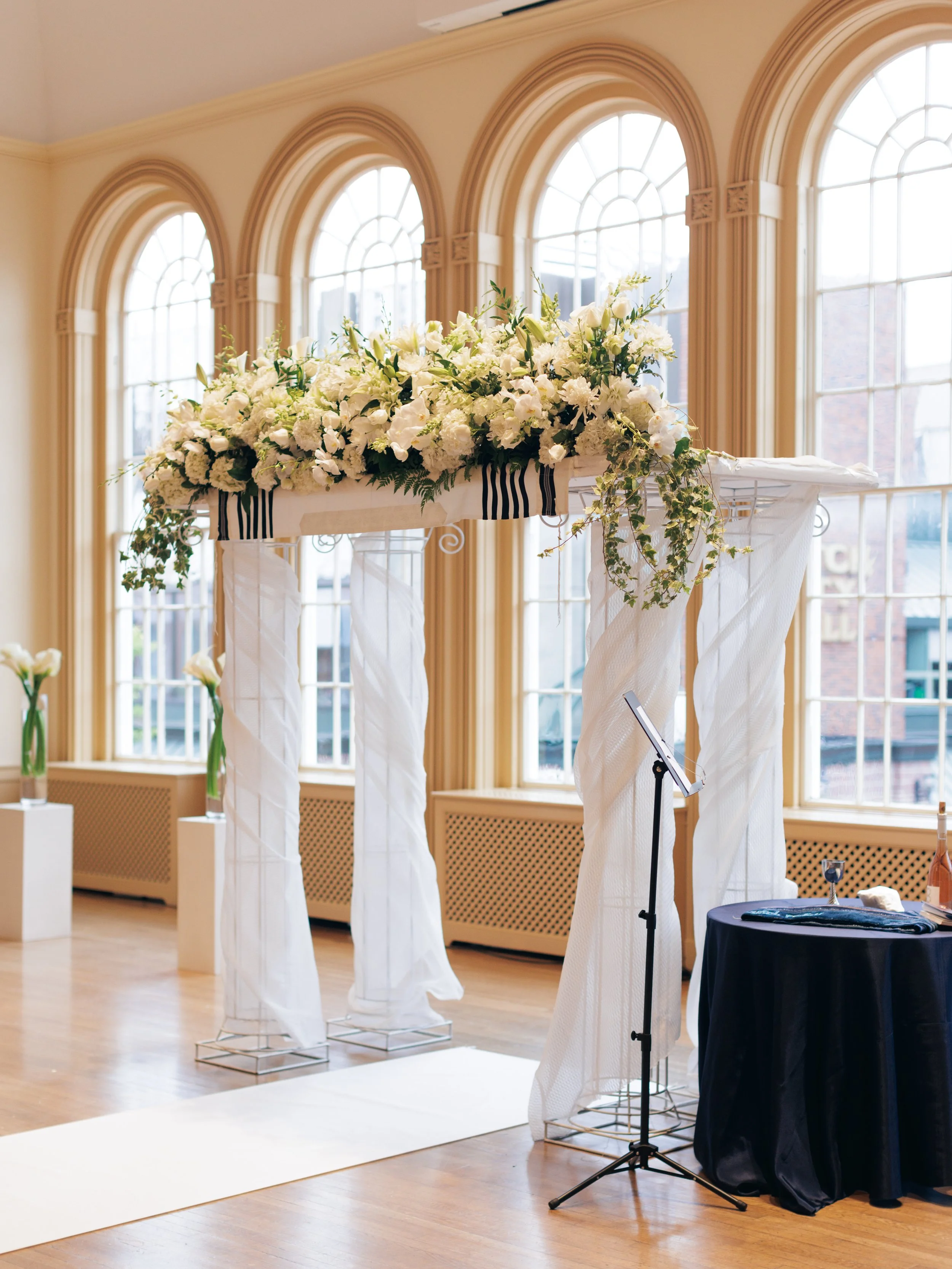An elegant wedding altar decorated with a lush arrangement of white flowers and greenery, set against large arched windows in a brightly lit room with wooden floors.