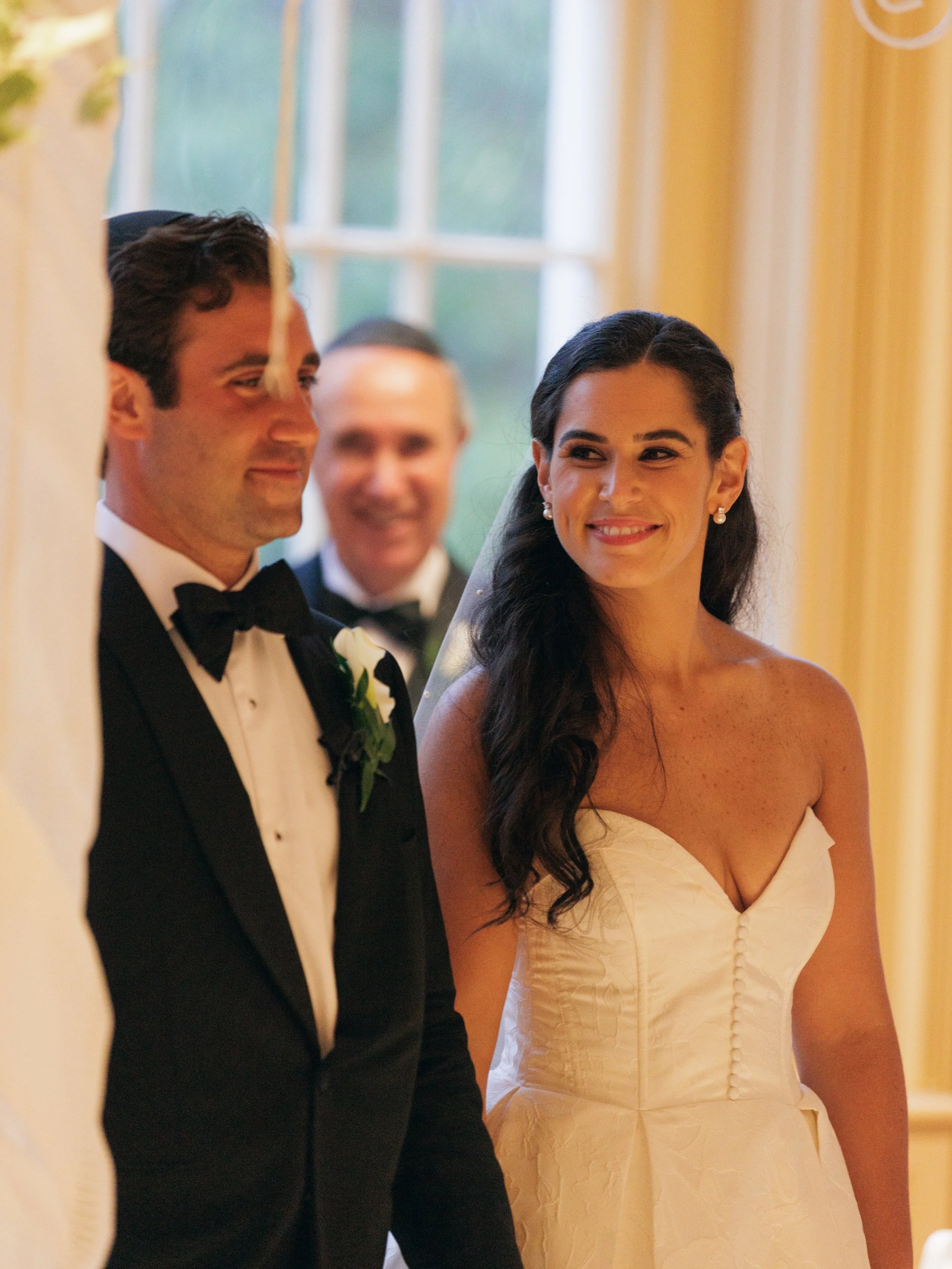 A bride and groom standing close together at their wedding, with the groom wearing a black tuxedo with a bow tie, and the bride in a strapless white wedding dress with dark wavy hair, smiling at each other.