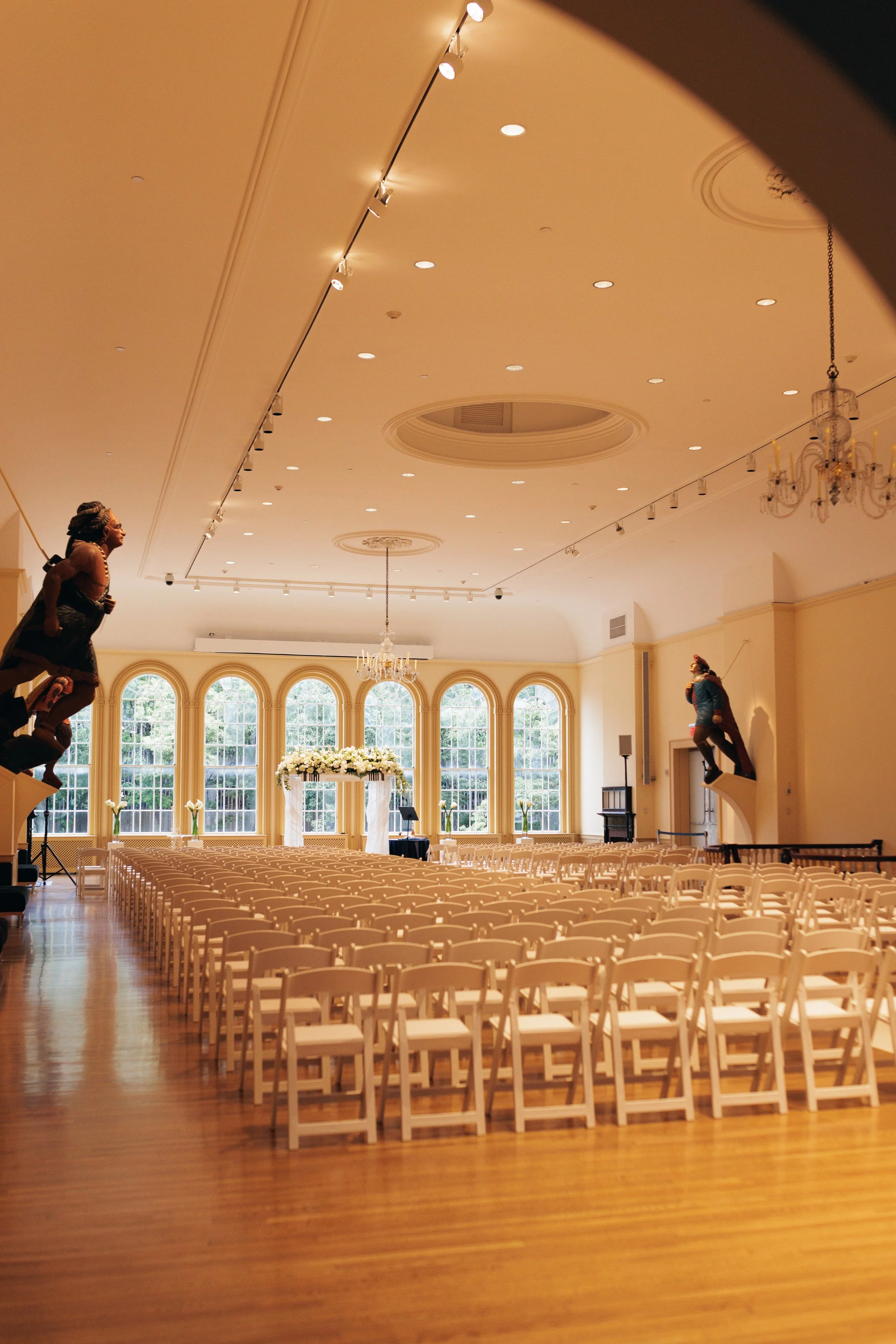 A large elegant hall with a high ceiling, chandeliers, and a row of arched windows letting in natural light. Rows of white chairs are set up facing a small platform decorated with flowers. The hall appears to be prepared for a wedding or large event.