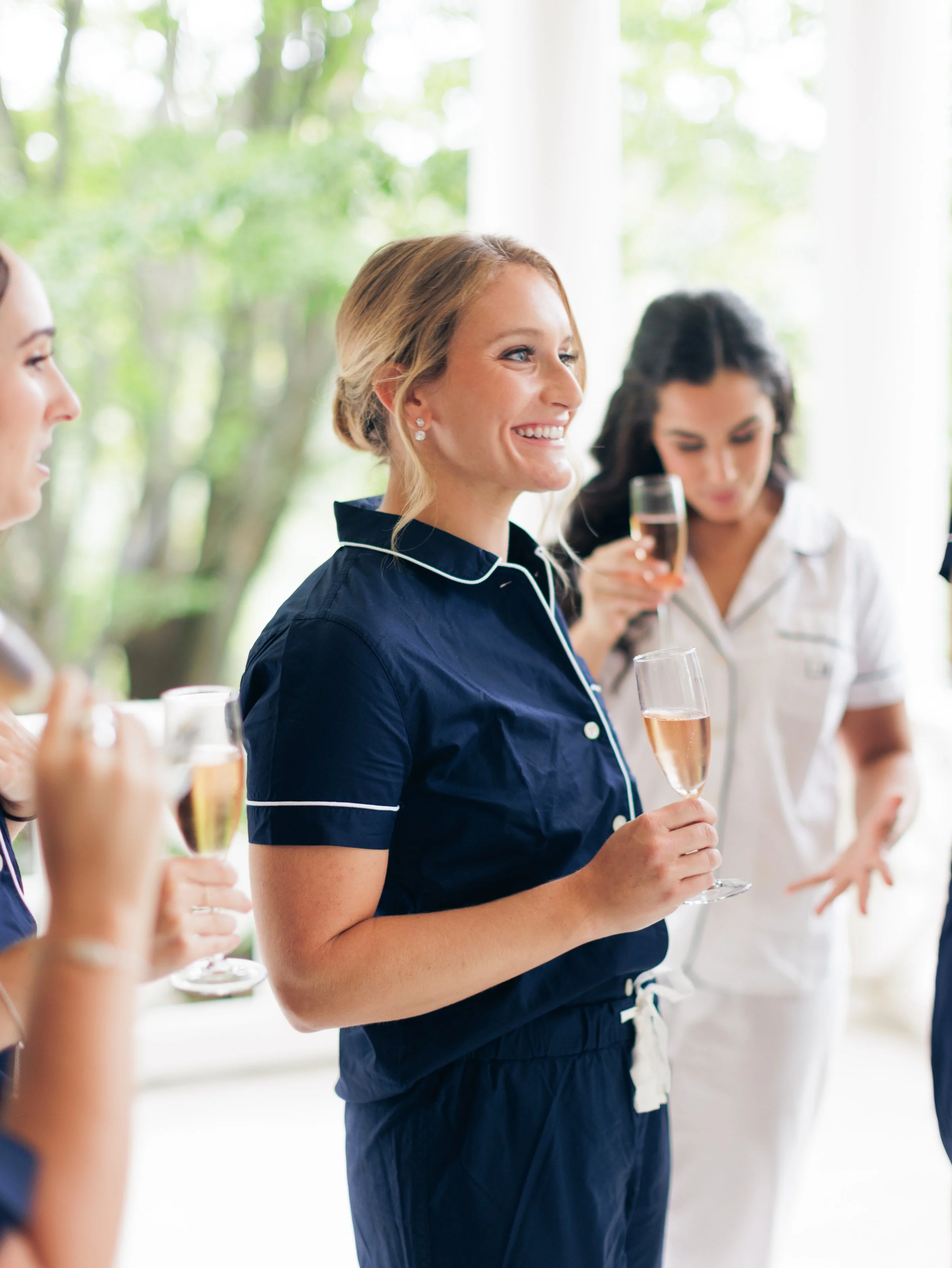 Women in pajamas holding glasses of champagne at a gathering, standing near a window with trees outside.