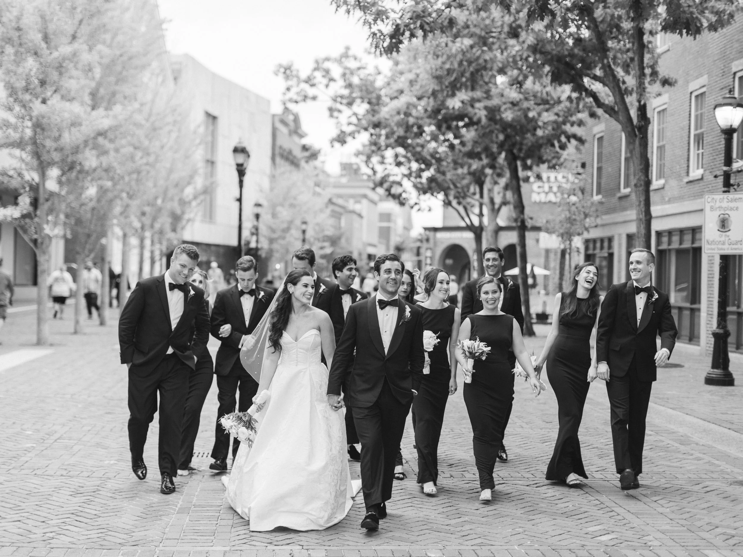 Black and white photo of a wedding procession with the bride and groom walking down a city street surrounded by friends in formal attire, enjoying a celebration.