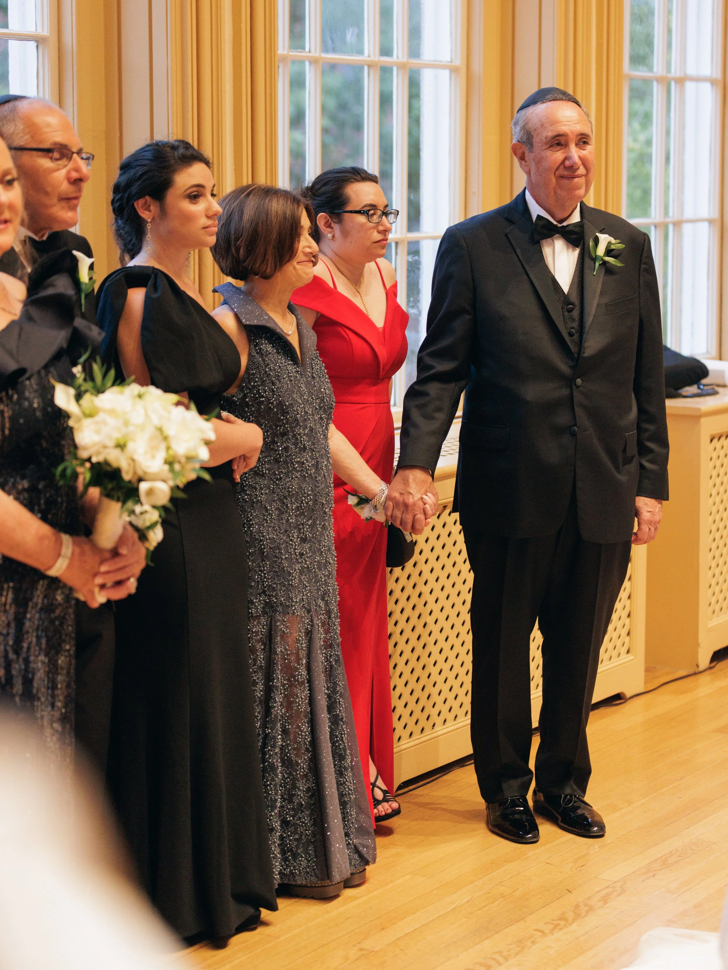 People standing in a line at a formal event, holding hands, dressed in tuxedos and elegant dresses, near large windows with yellow frames.