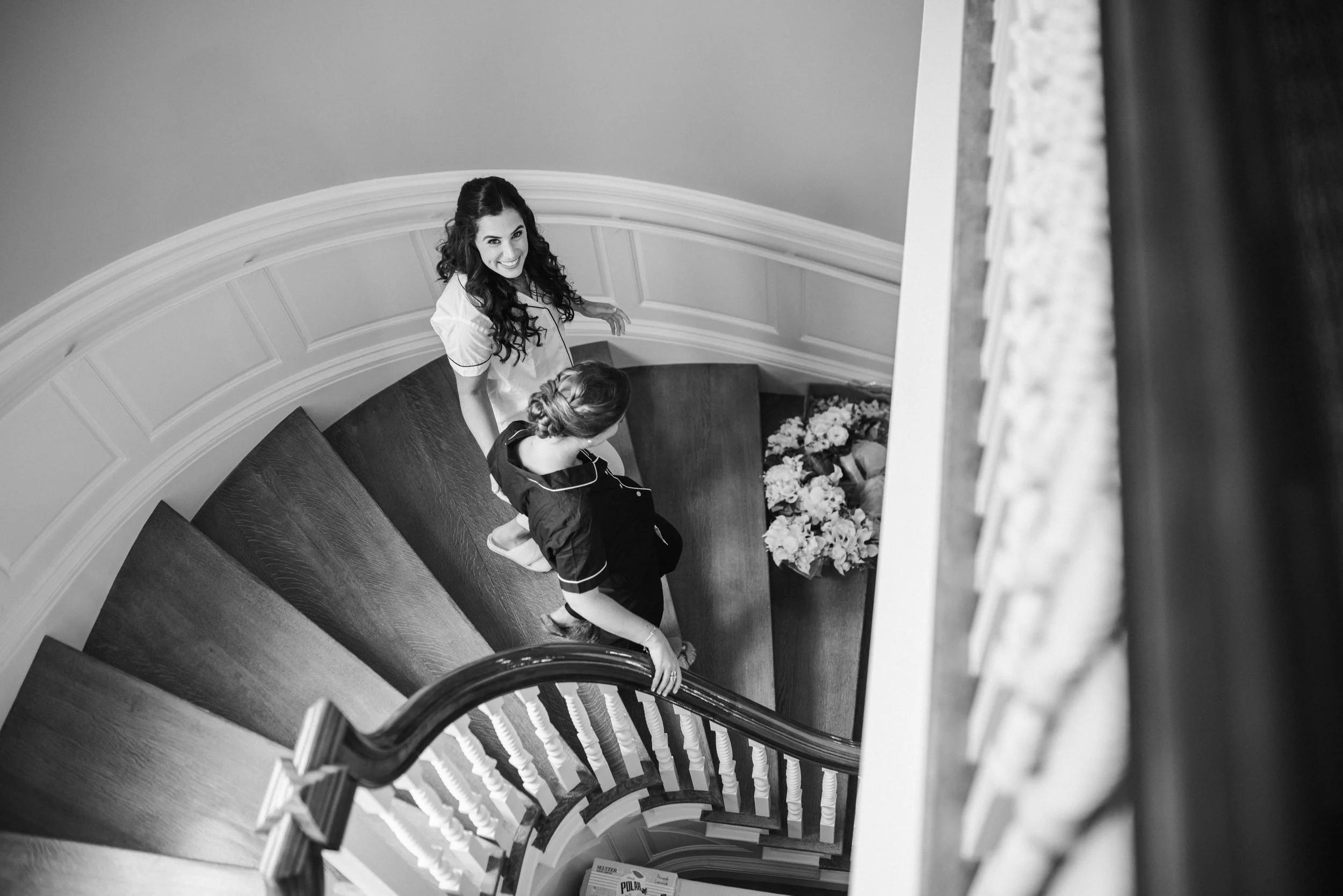 Black-and-white photo taken from the top of a curved staircase with a woman smiling and looking up at the camera, standing on the stairs, with another woman sitting on the stairs with her back to the camera, in front of a wall with a flower arrangeme