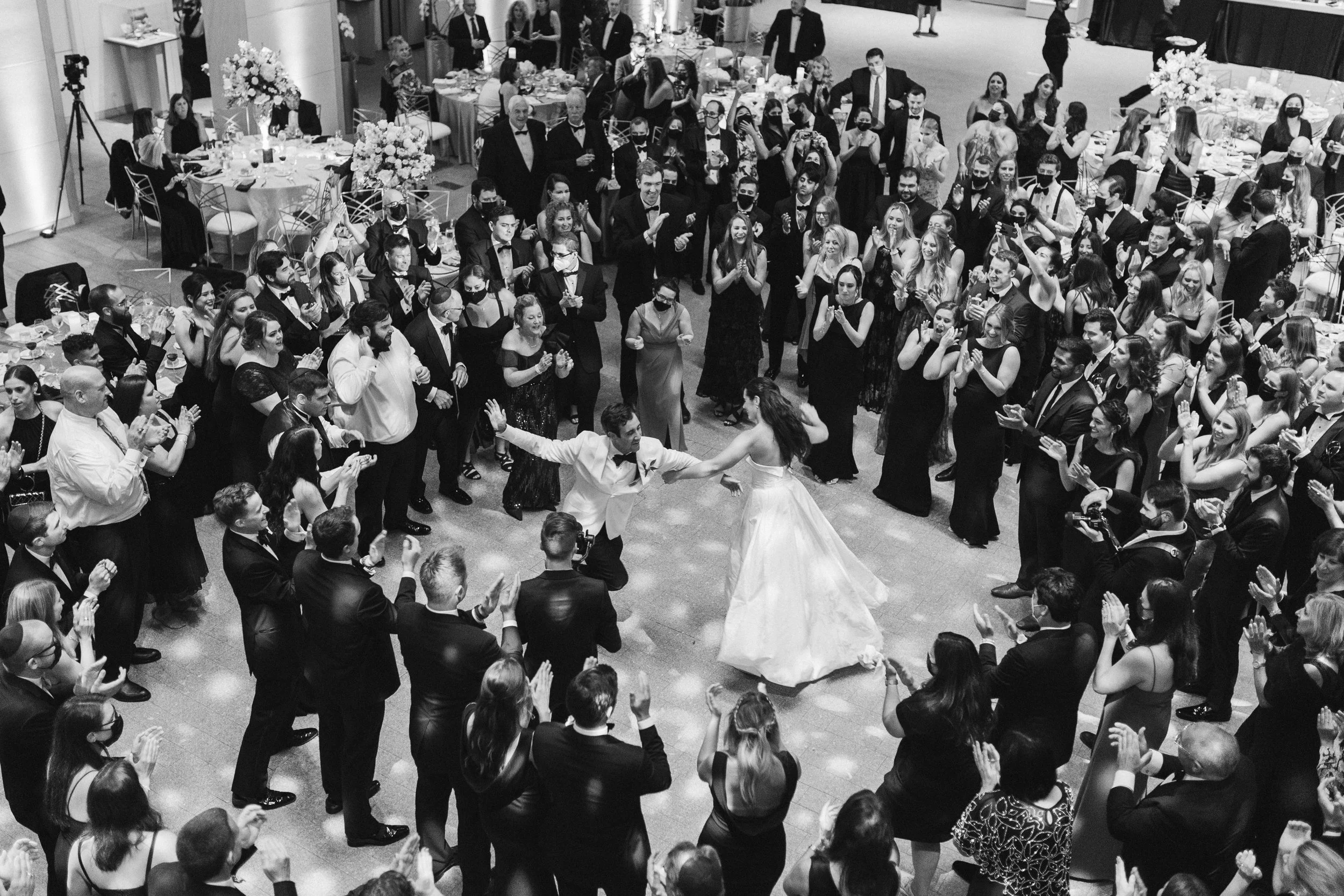 A black-and-white photo of a large indoor party with people in formal attire. A couple is dancing in the center, surrounded by guests clapping and watching. The guests are wearing suits and dresses, with some wearing masks. The tables are decorated w
