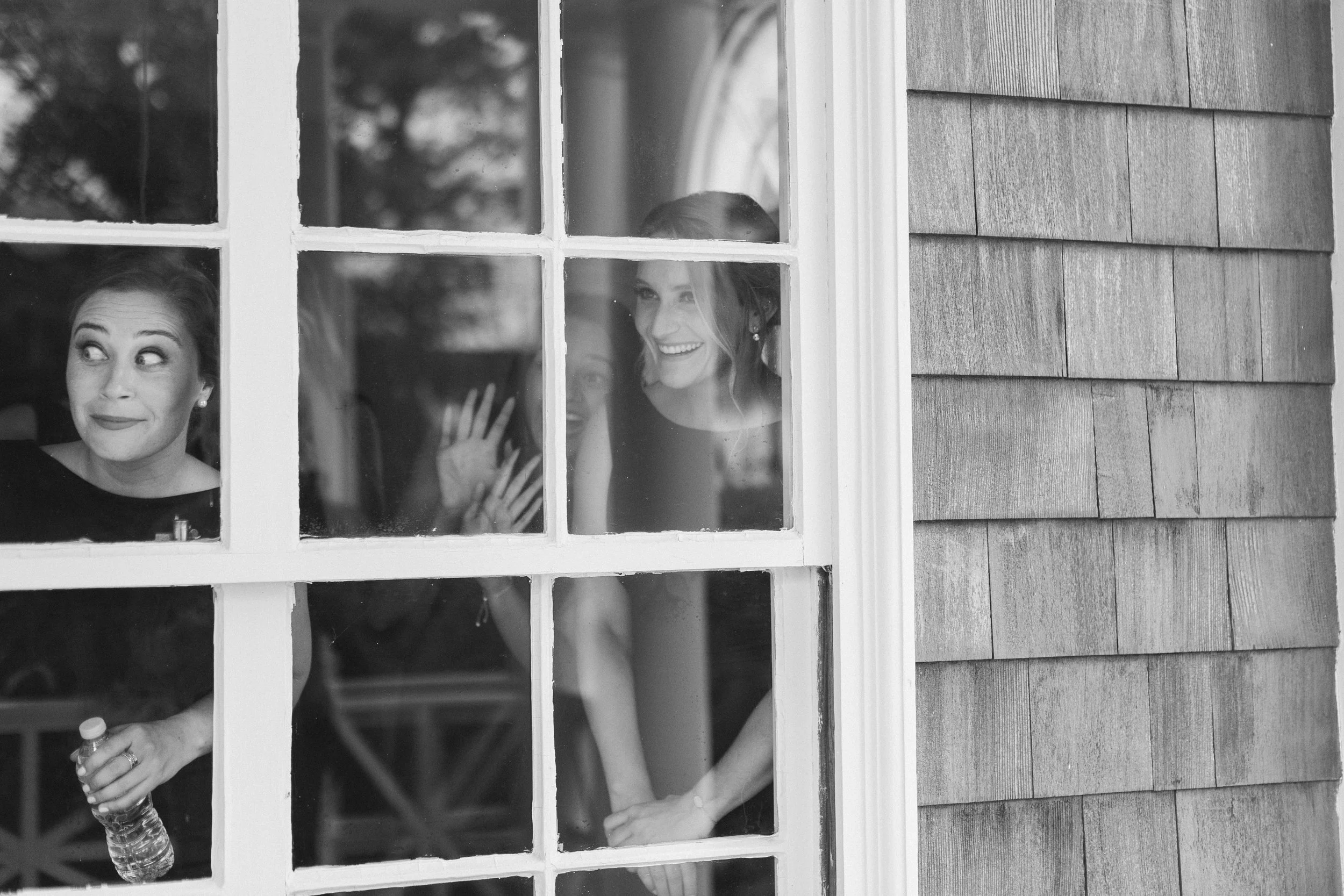 Three women looking out a window, smiling and talking, with one holding a water bottle.