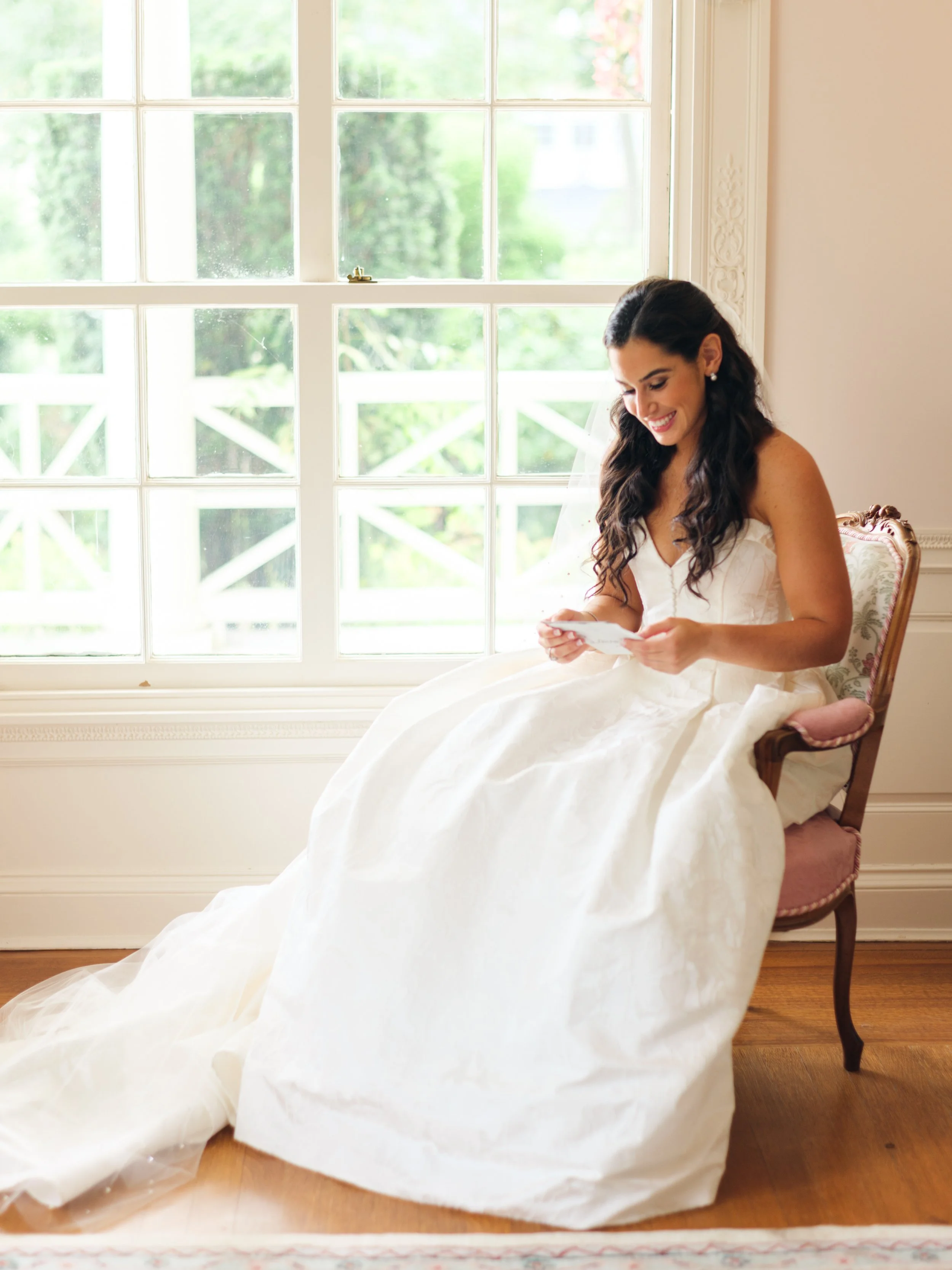 A woman in a wedding dress sitting in a vintage chair near a large window, smiling while looking at something in her hands.