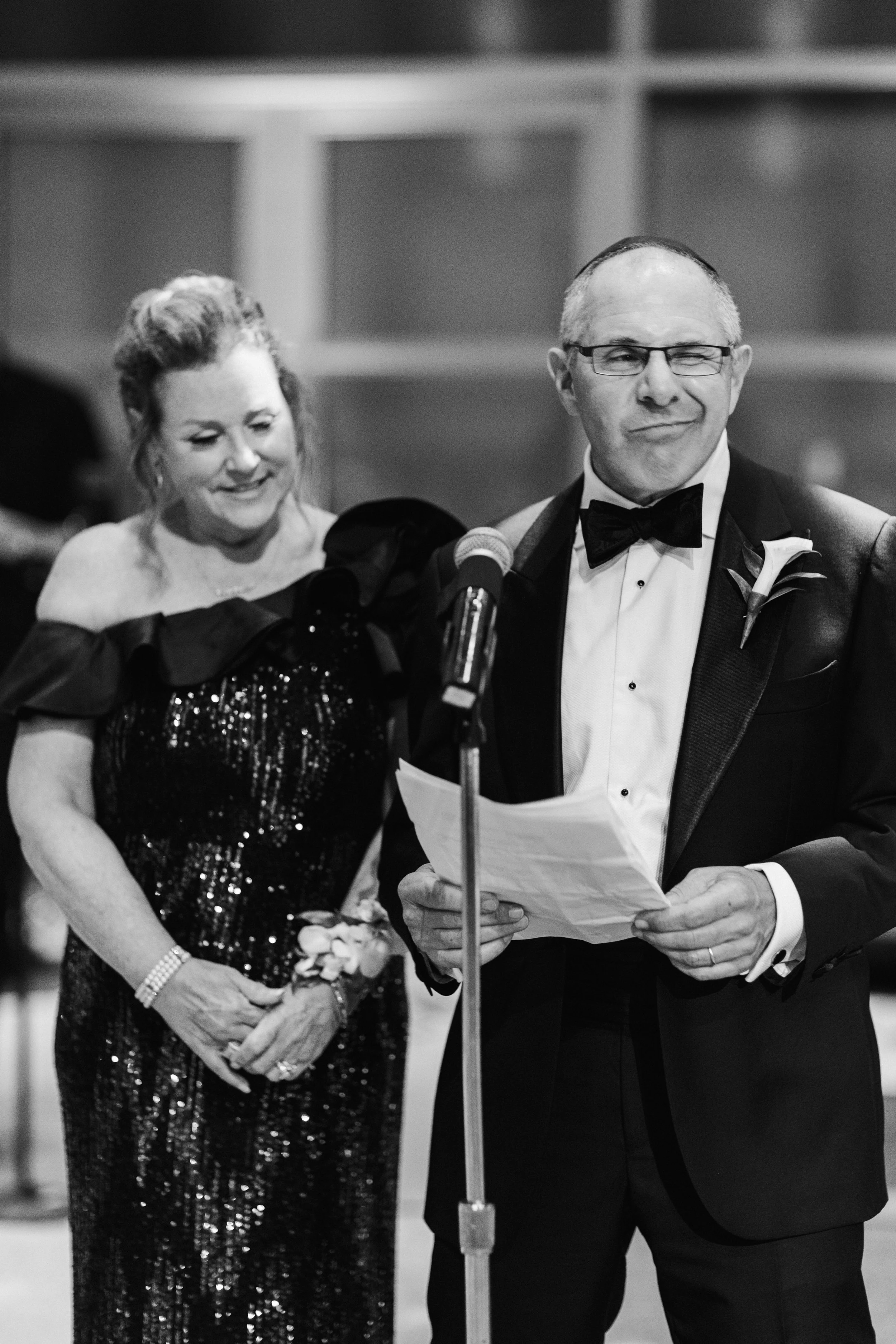 A black and white photo of a man and woman at a formal event. The woman, dressed in a sequined gown, is smiling and holding a corsage, while the man, wearing a tuxedo with glasses and a bow tie, is speaking at a microphone and holding papers.