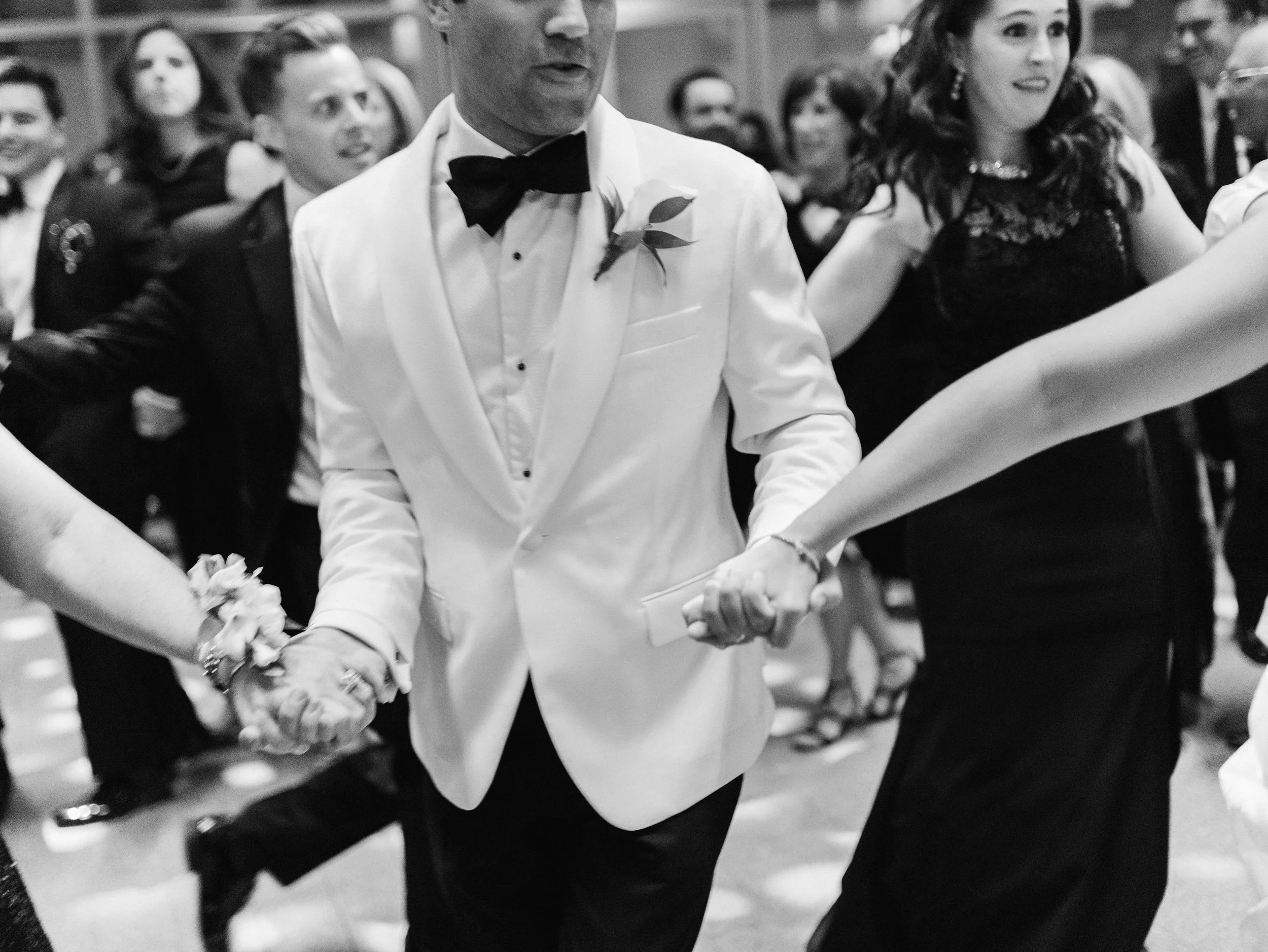 People are dancing at a formal event, with a man in a tuxedo holding hands with a woman in a dress, among other dancing couples in the background.
