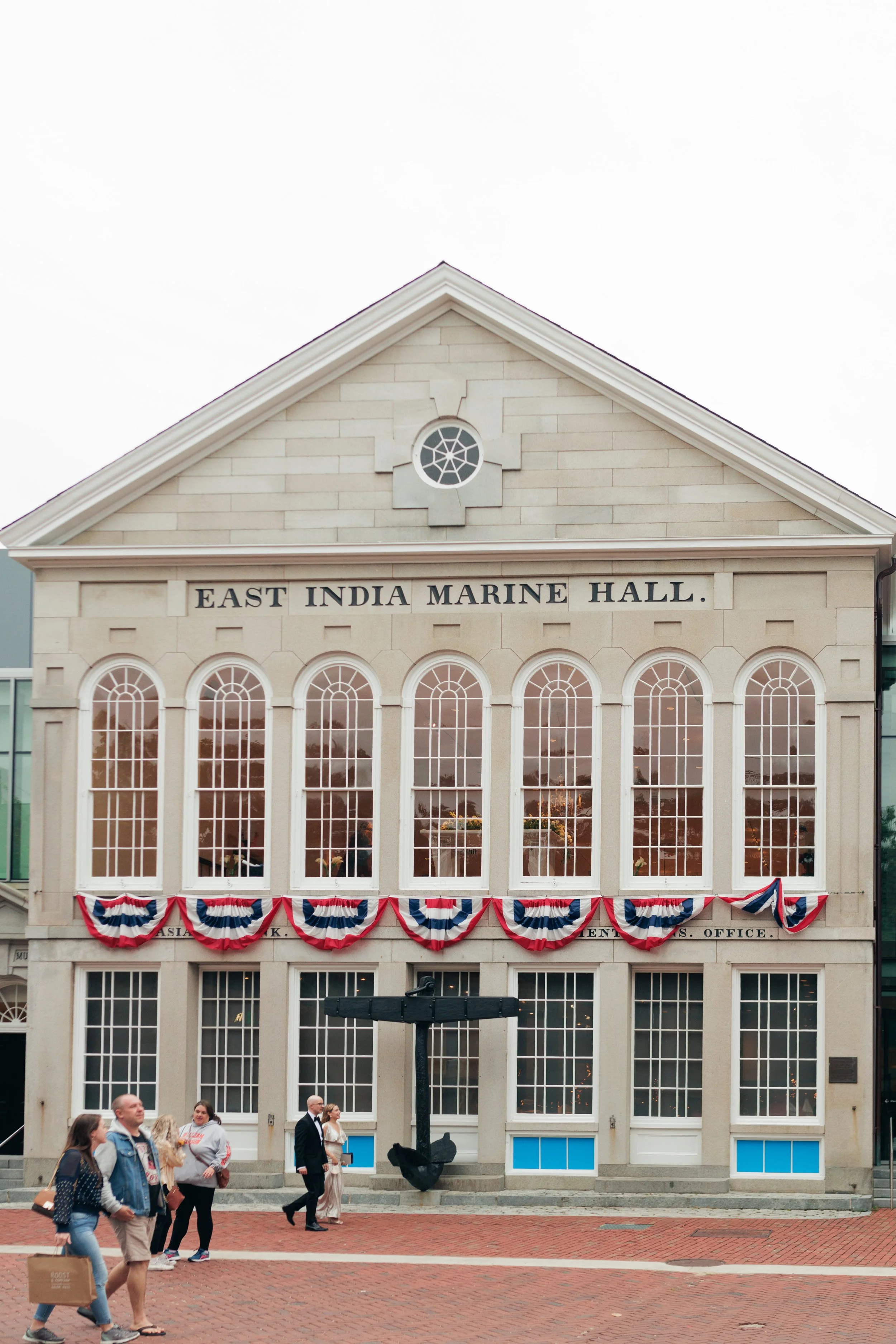 The East India Marine Hall building with patriotic banners, a protruding anchor sculpture, and people walking in front of it on a brick-paved area