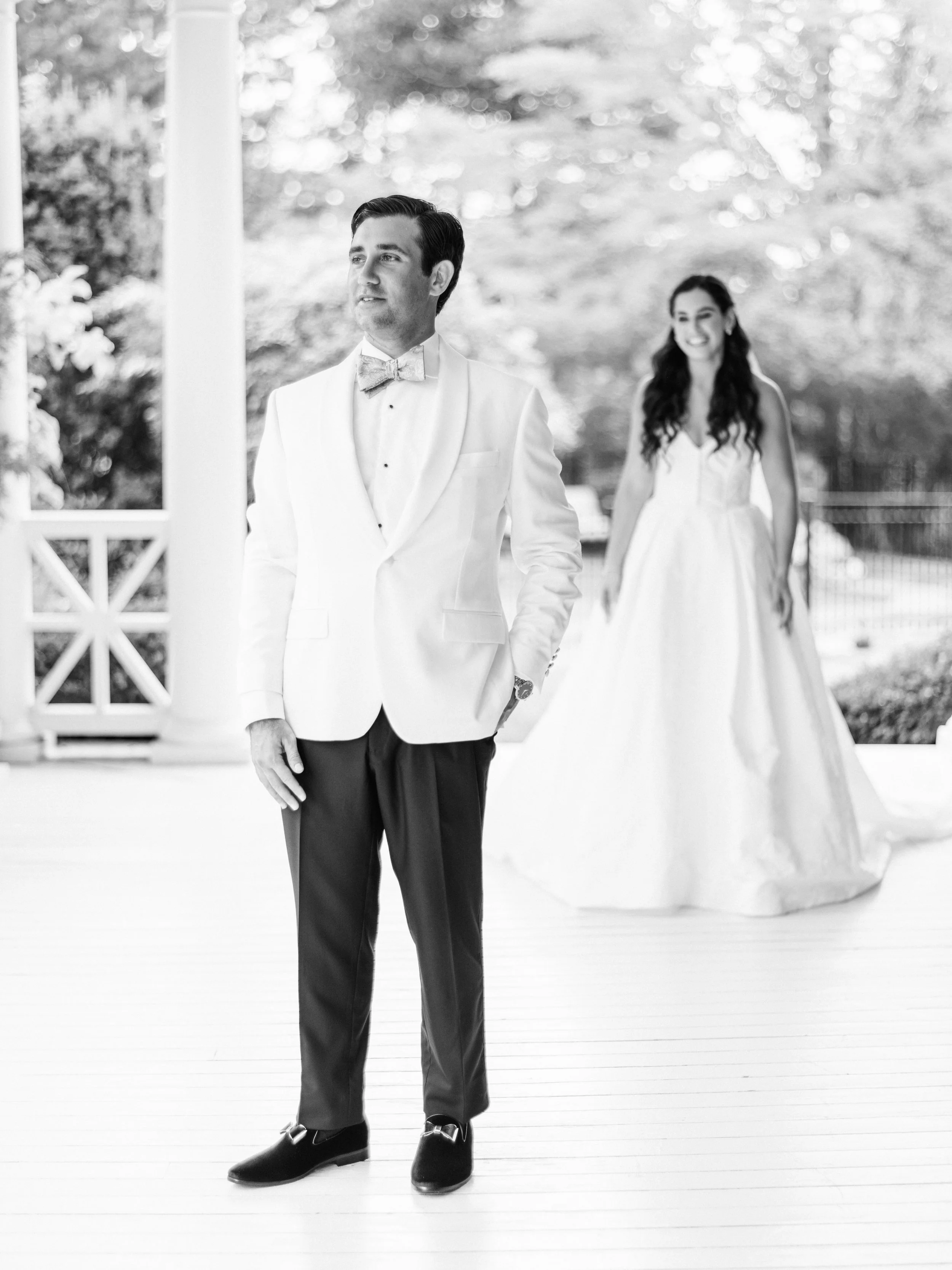 A man dressed in a white tuxedo with a bow tie, standing outdoors on a wooden deck. A woman in a white dress is in the background, smiling. The scene appears to be a wedding or formal event.