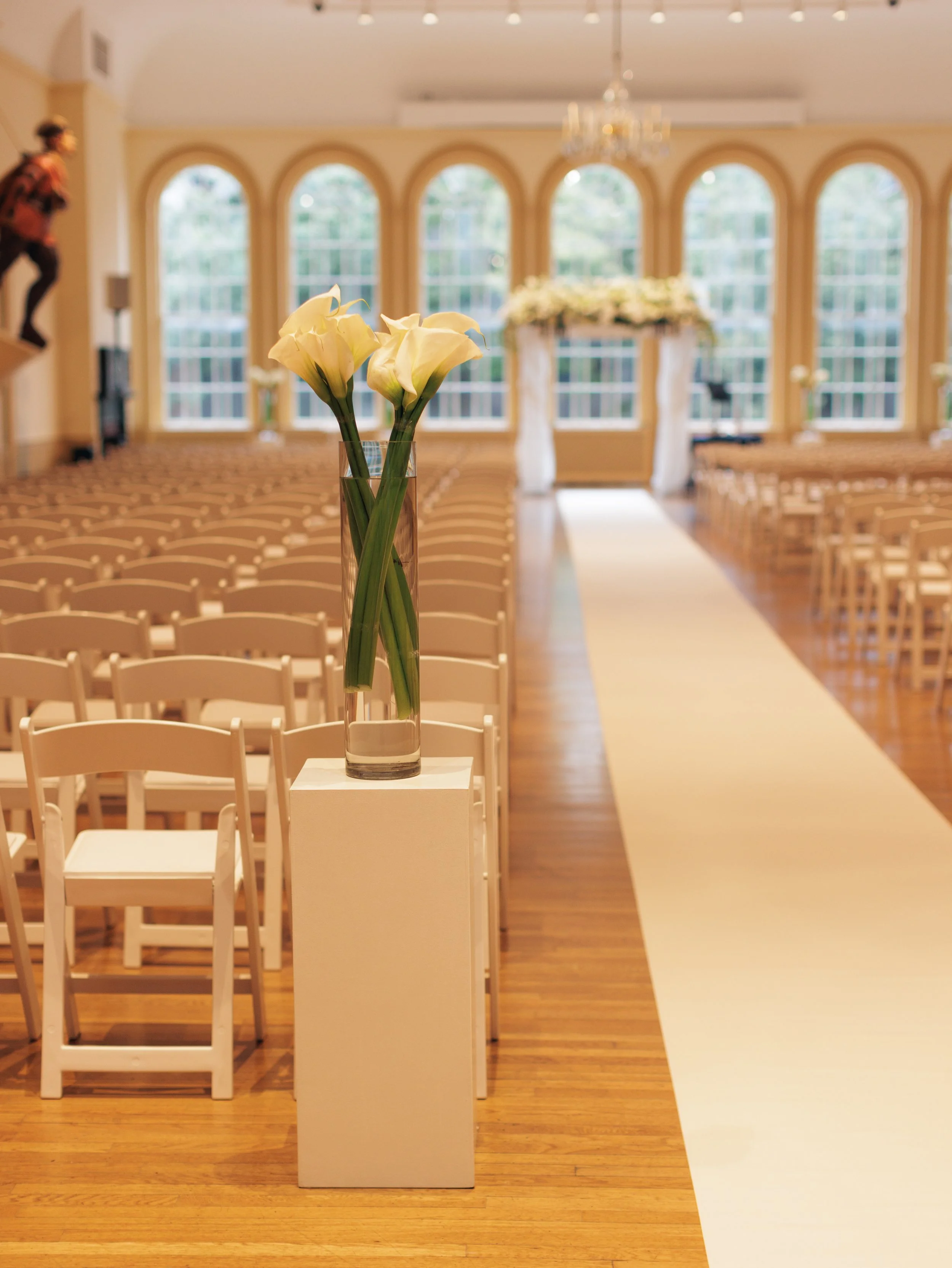 A tall glass vase with white calla lilies on a white pedestal in front of empty chairs arranged in rows, a white aisle runner, and a decorated altar at the front of a well-lit room with large arched windows.