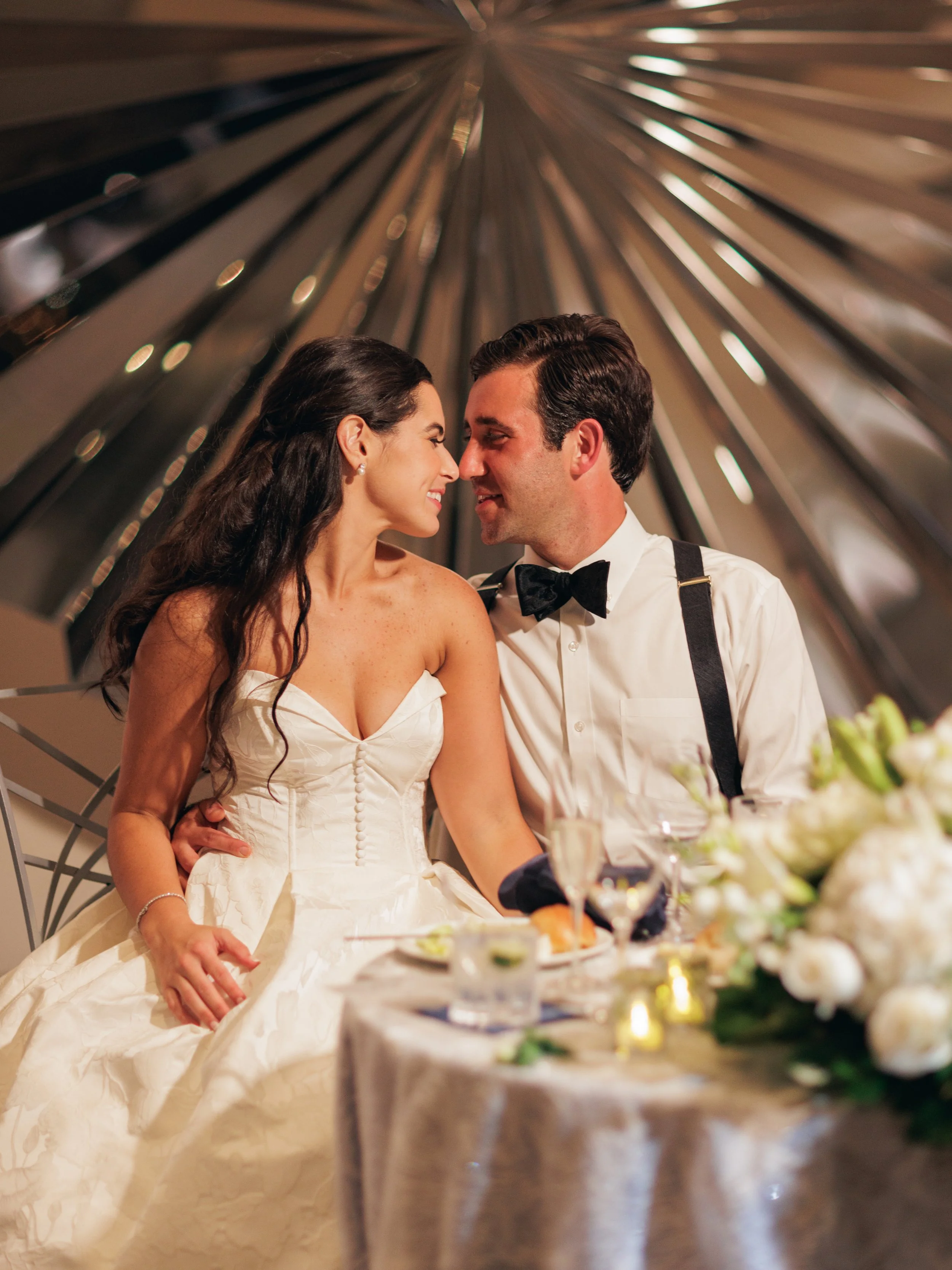 A bride and groom on their wedding day, sitting close together at a reception table with their foreheads and noses touching, smiling at each other with a warm expression. The bride wears a strapless white wedding gown and the groom wears a tuxedo wit