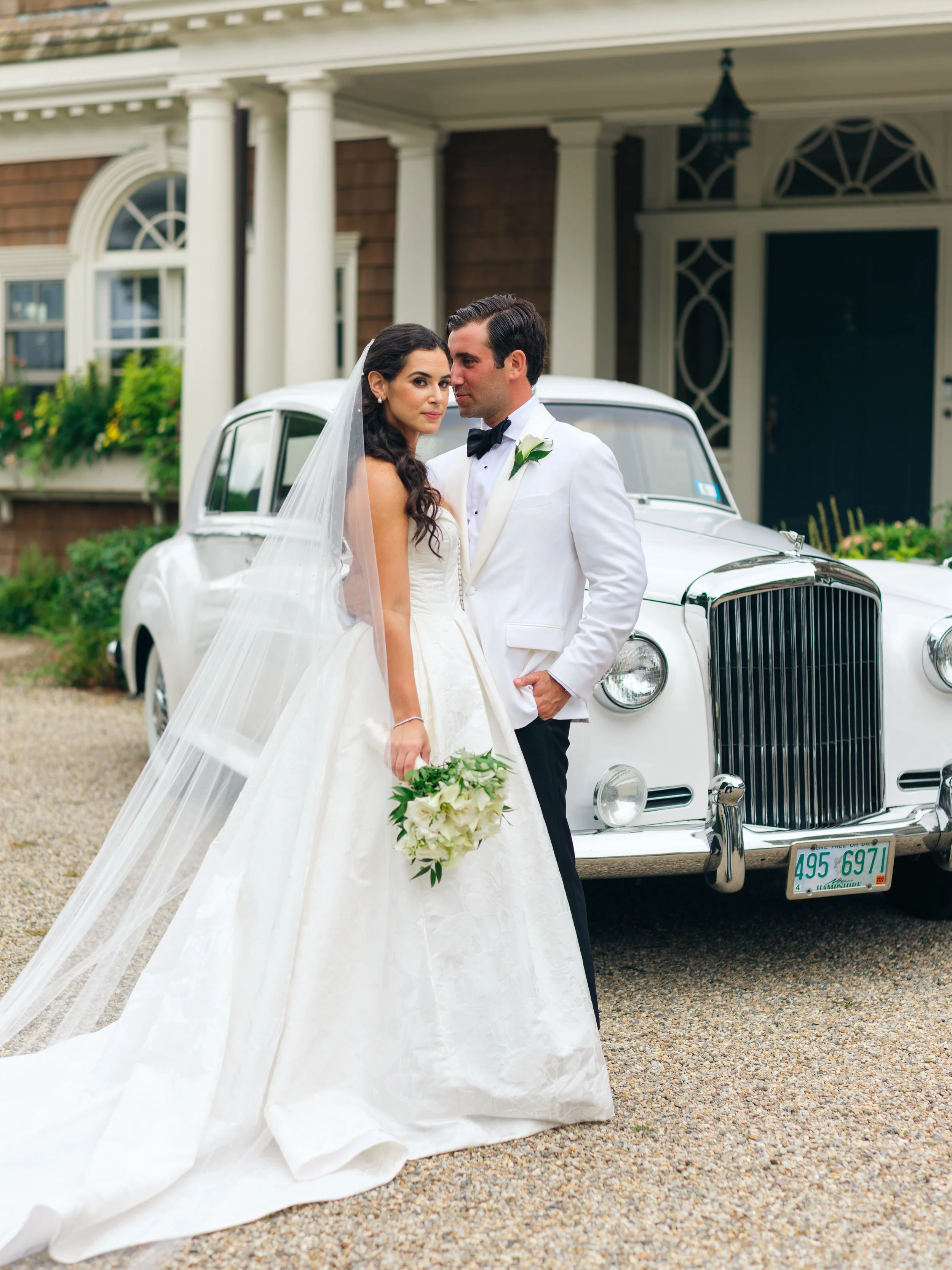 A bride and groom in wedding attire standing in front of a white vintage car, with a large house in the background.