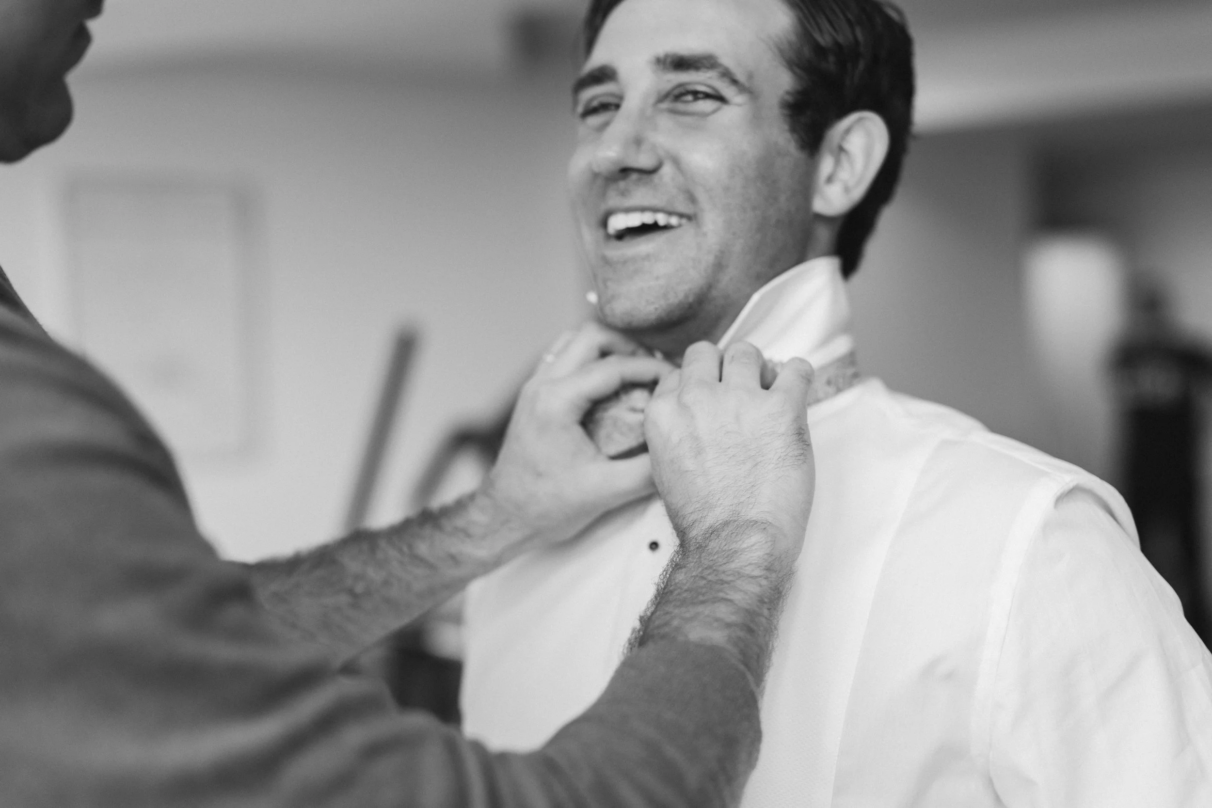 A man in a tuxedo is being assisted by another person in a white shirt to adjust his bowtie, smiling.
