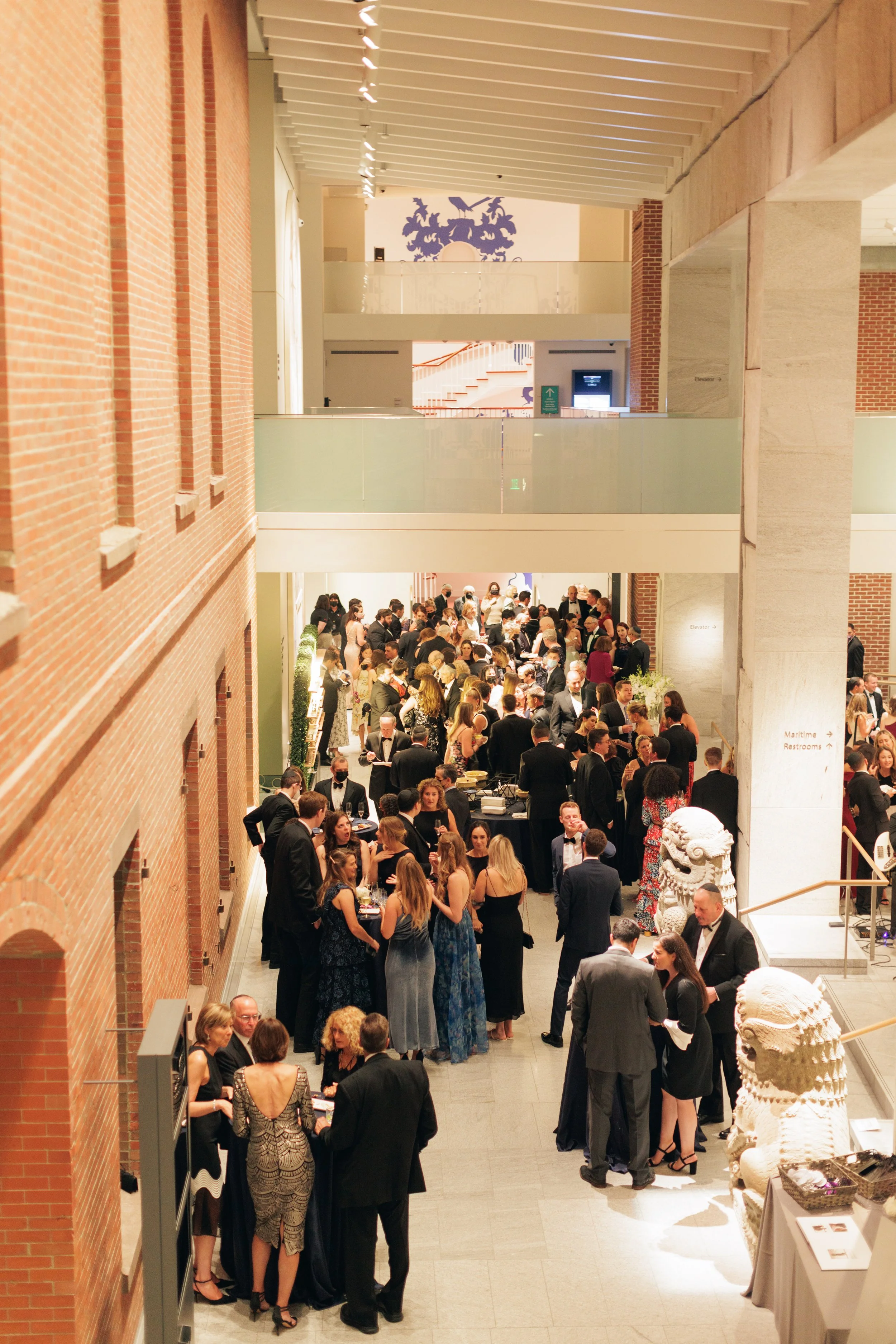 A large indoor gathering or reception with many people dressed in formal attire, including tuxedos and evening gowns, in a spacious venue with brick walls, modern decor, and sculptures, likely a museum or event hall.