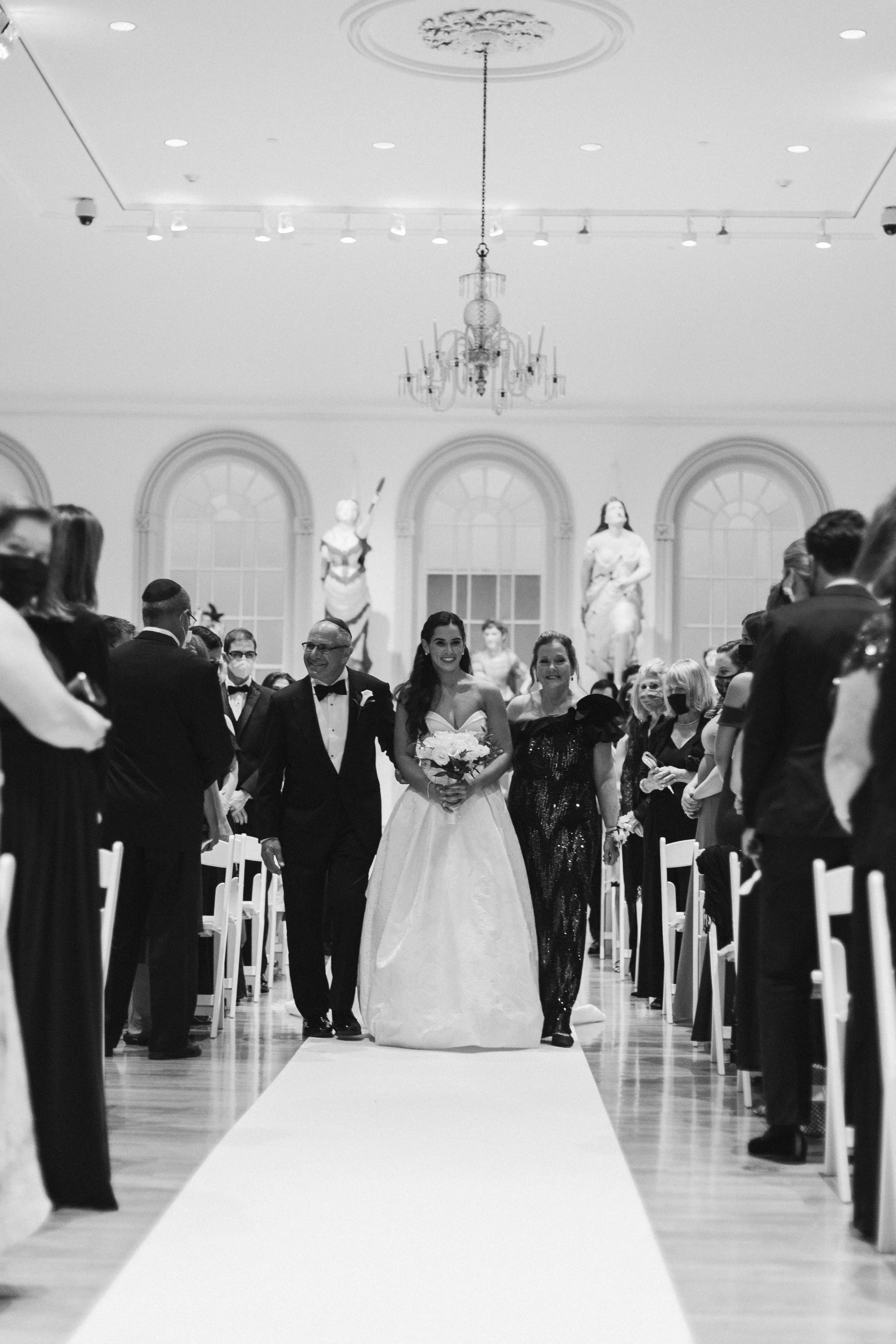 A bride in a white wedding gown walking down the aisle at a wedding ceremony, flanked by guests on both sides, inside an elegant hall with statues and large windows.