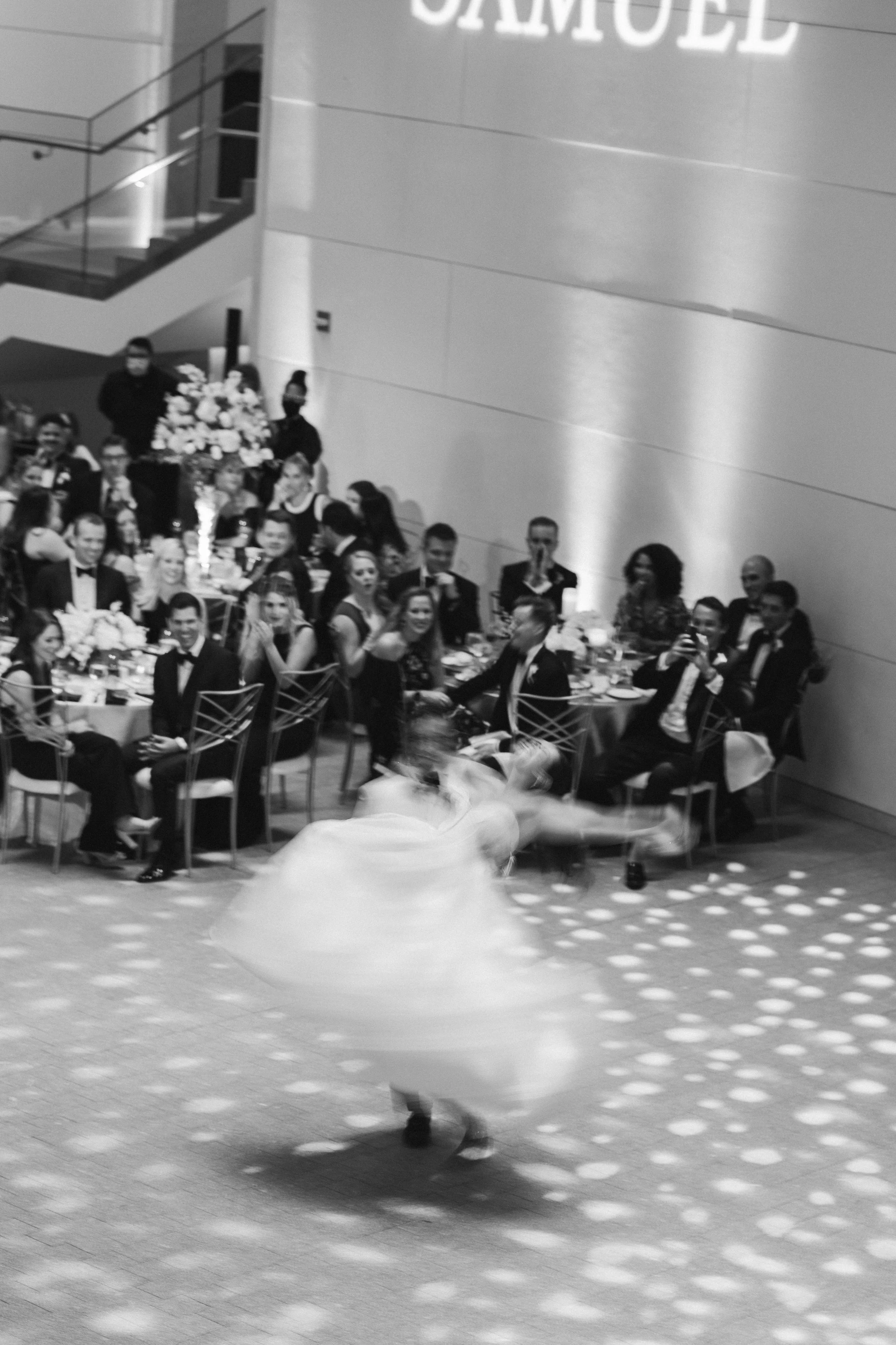 People dancing in a large hall with tables of formally dressed guests watching, some clapping, during a celebration or event.
