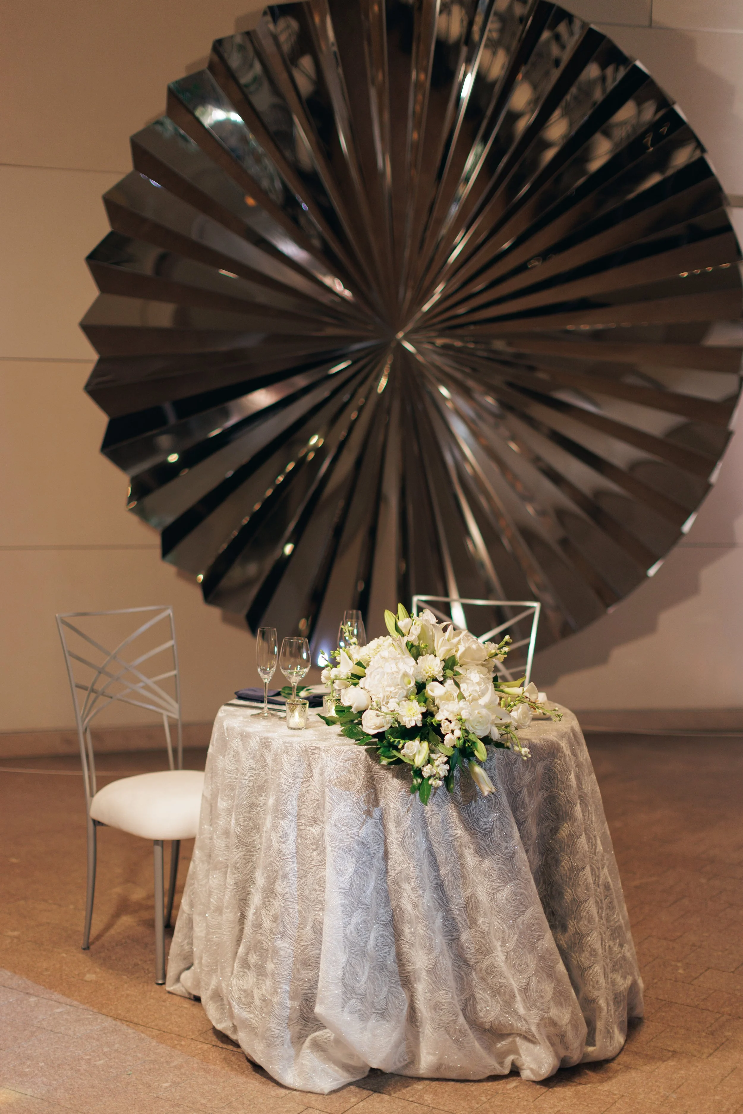 A table decorated with white and green floral centerpiece, surrounded by clear glasses, set for a celebration or wedding, against a backdrop with a large black fan-shaped wall decoration.
