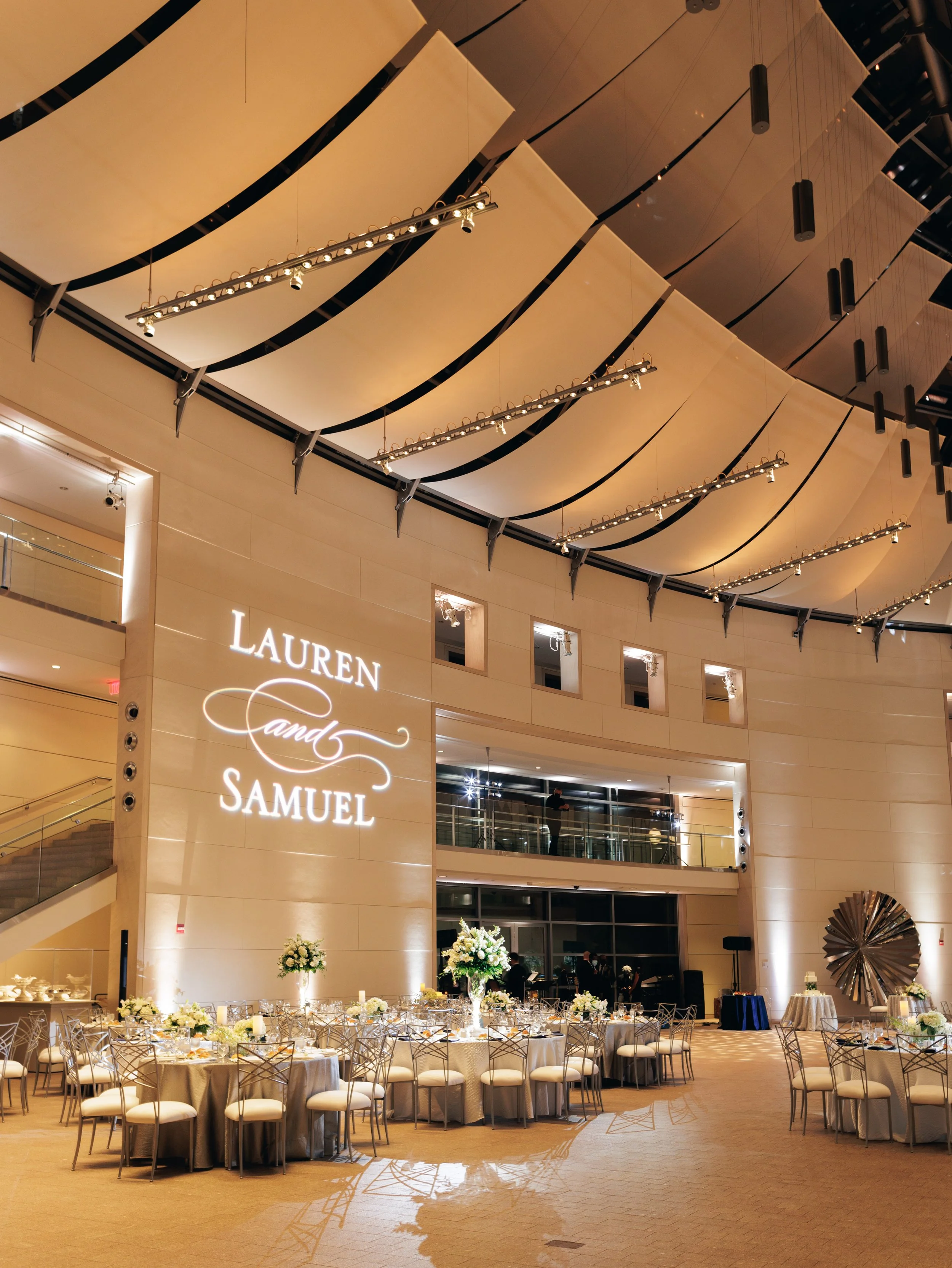 Elegant banquet hall decorated for a formal event with round tables covered in white linens, surrounded by chairs, adorned with floral centerpieces, and illuminated by decorative lighting, with a sign reading 'Lauren Candy Samuel' and floral arrangem