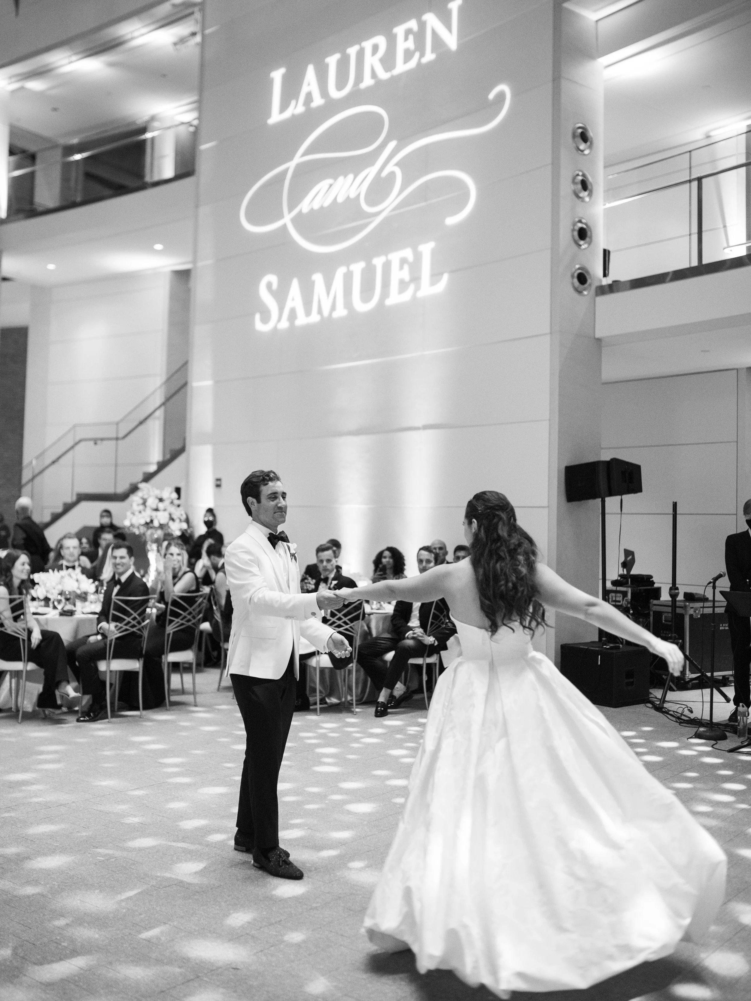 A black and white photo of a wedding reception inside a modern venue. A couple is dancing; the groom is in a tuxedo with a bow tie, and the bride is in a voluminous wedding dress, holding one arm out. Guests are seated at tables in the background, an