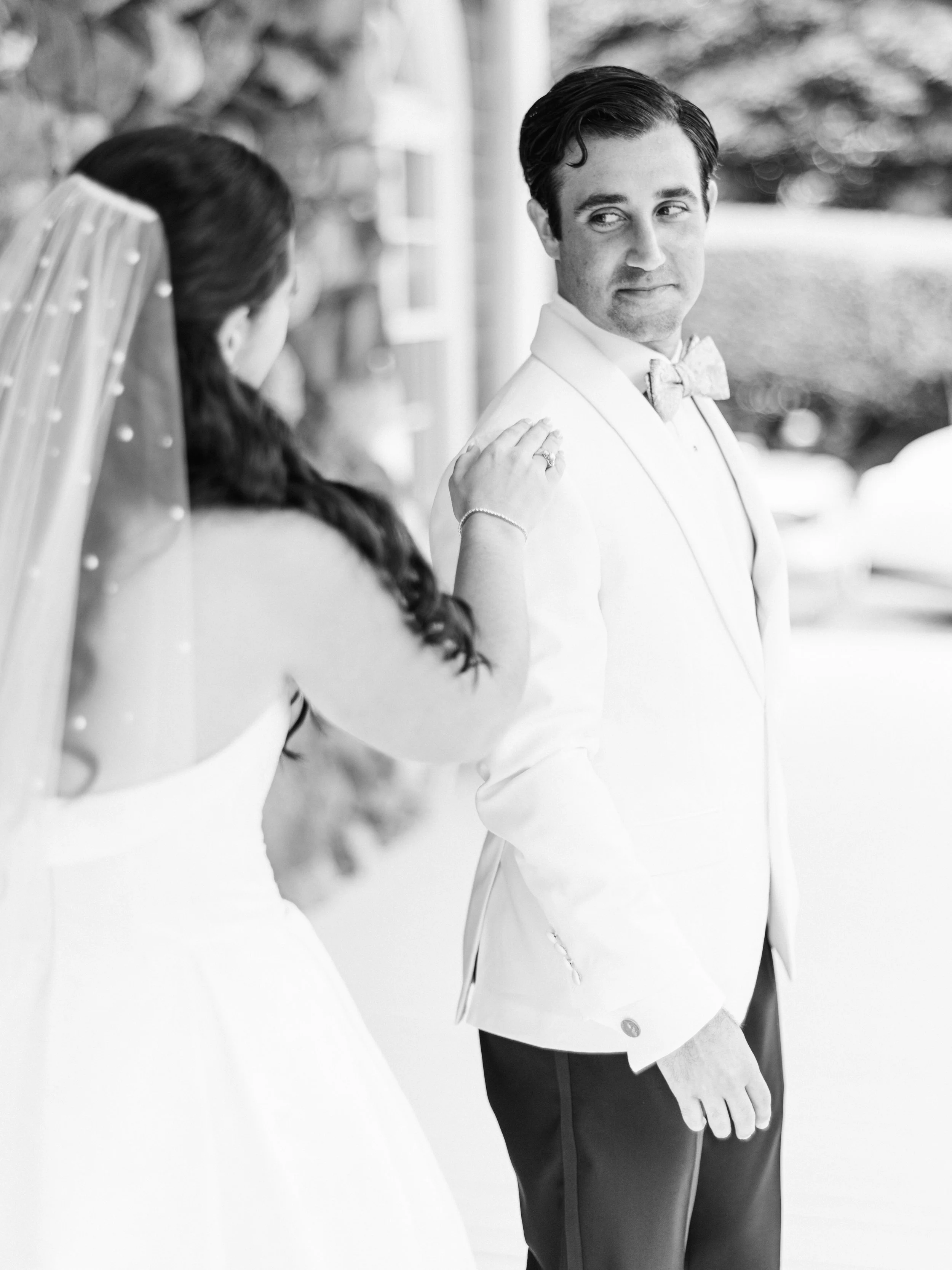 A black and white photo of a groom in a white tuxedo with a bow tie, smiling and looking to the side, standing outdoors with a blurred background. Partially visible is a woman with braided hair in a white dress and veil, touching the groom's shoulder