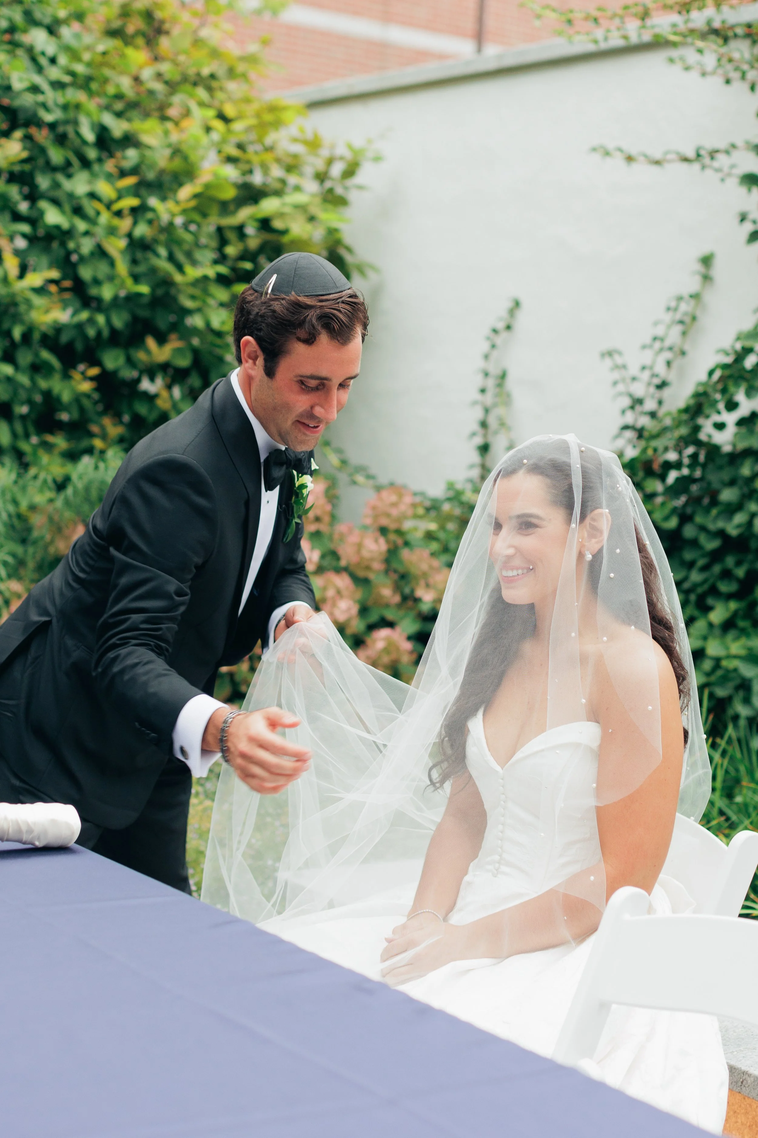 A groom in a black tuxedo helps a bride in a white wedding dress with her veil outdoors during a wedding ceremony. The bride is smiling, and the scene is surrounded by greenery and a white wall.