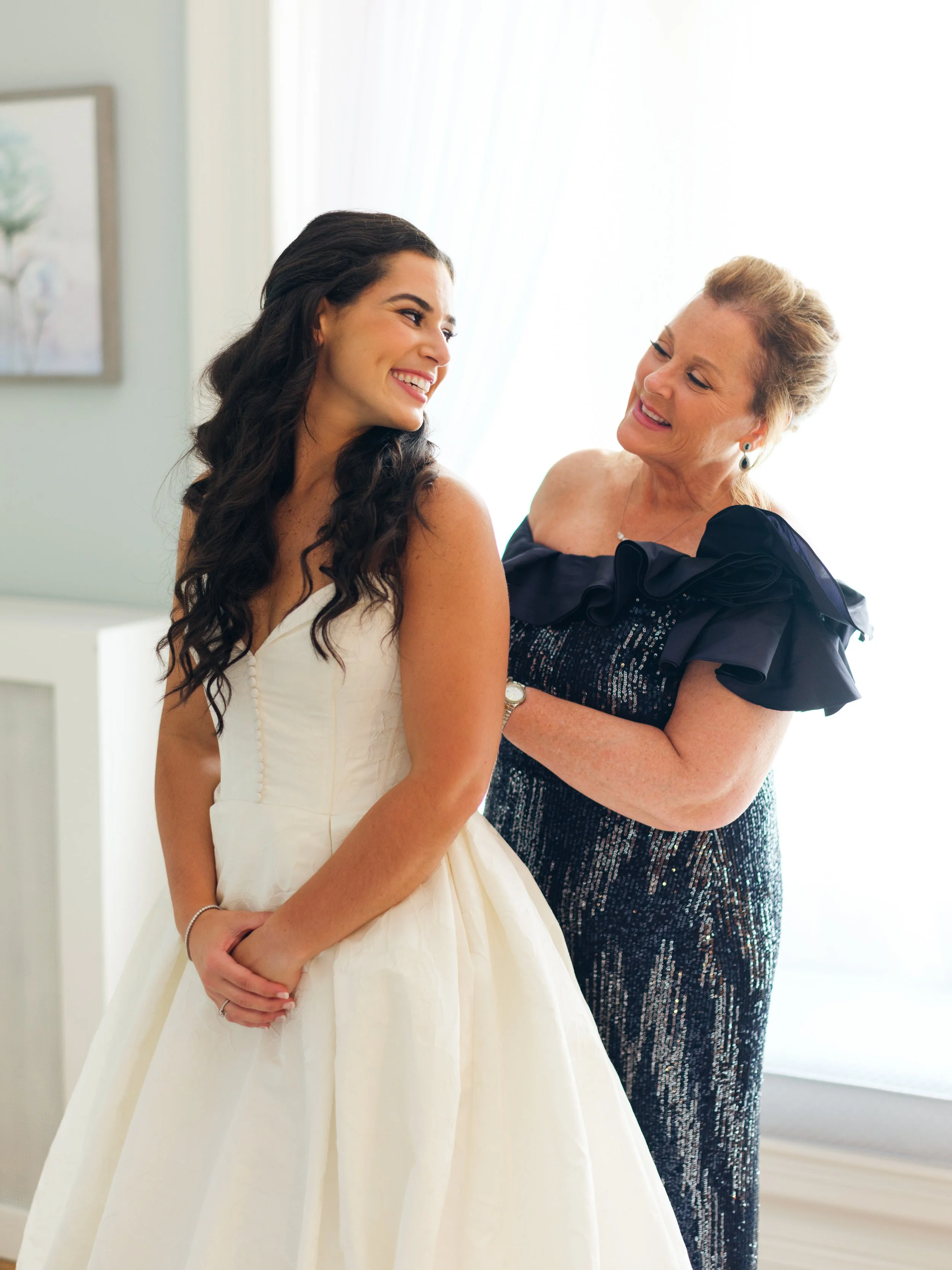 A young woman in a white dress smiling and looking at an older woman in a black dress with sequins, who is also smiling and looking at her.