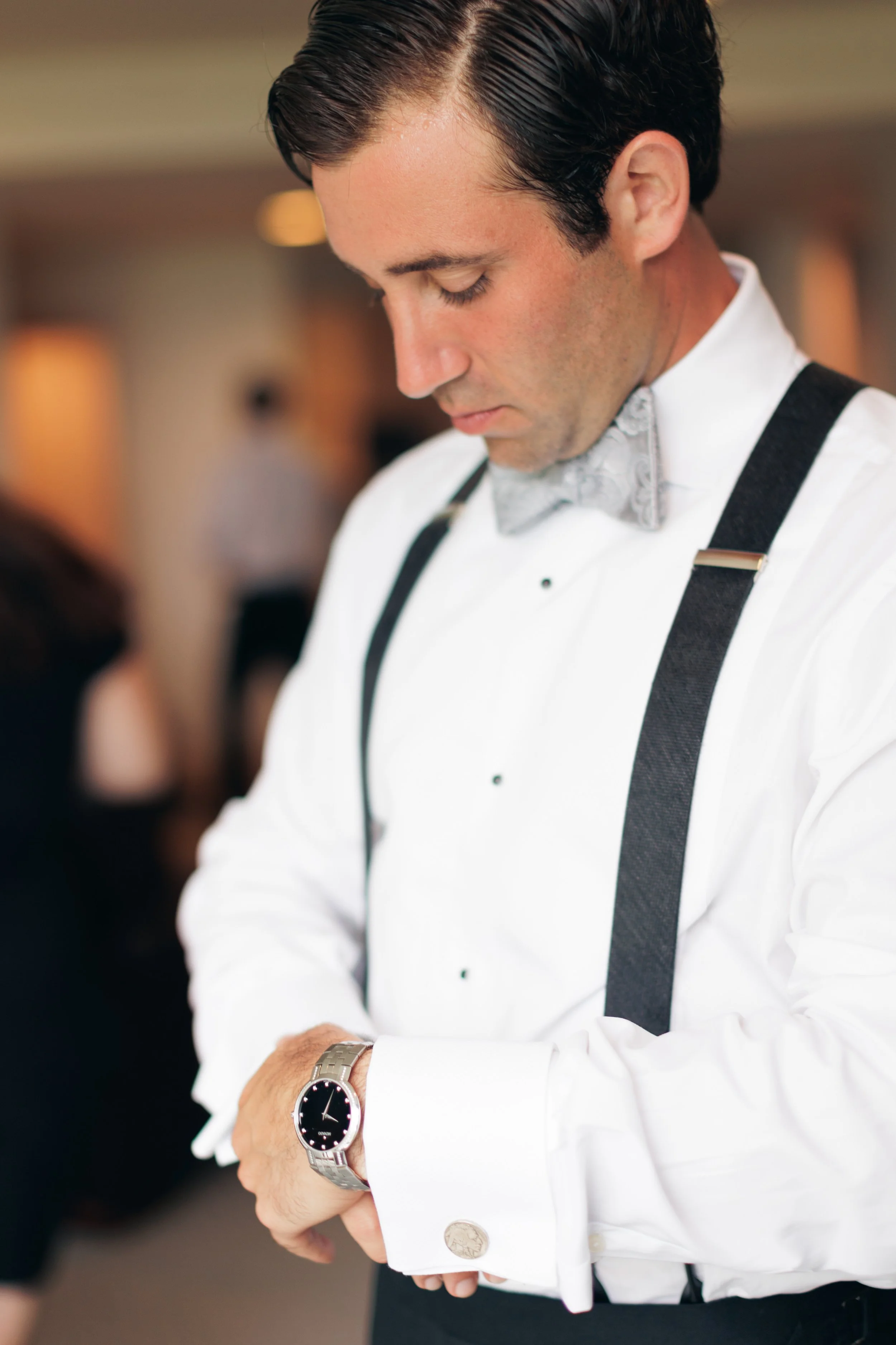 A man dressed in a white shirt with suspenders, wearing a silver watch, looking down as he adjusts his sleeve or watches his watch.