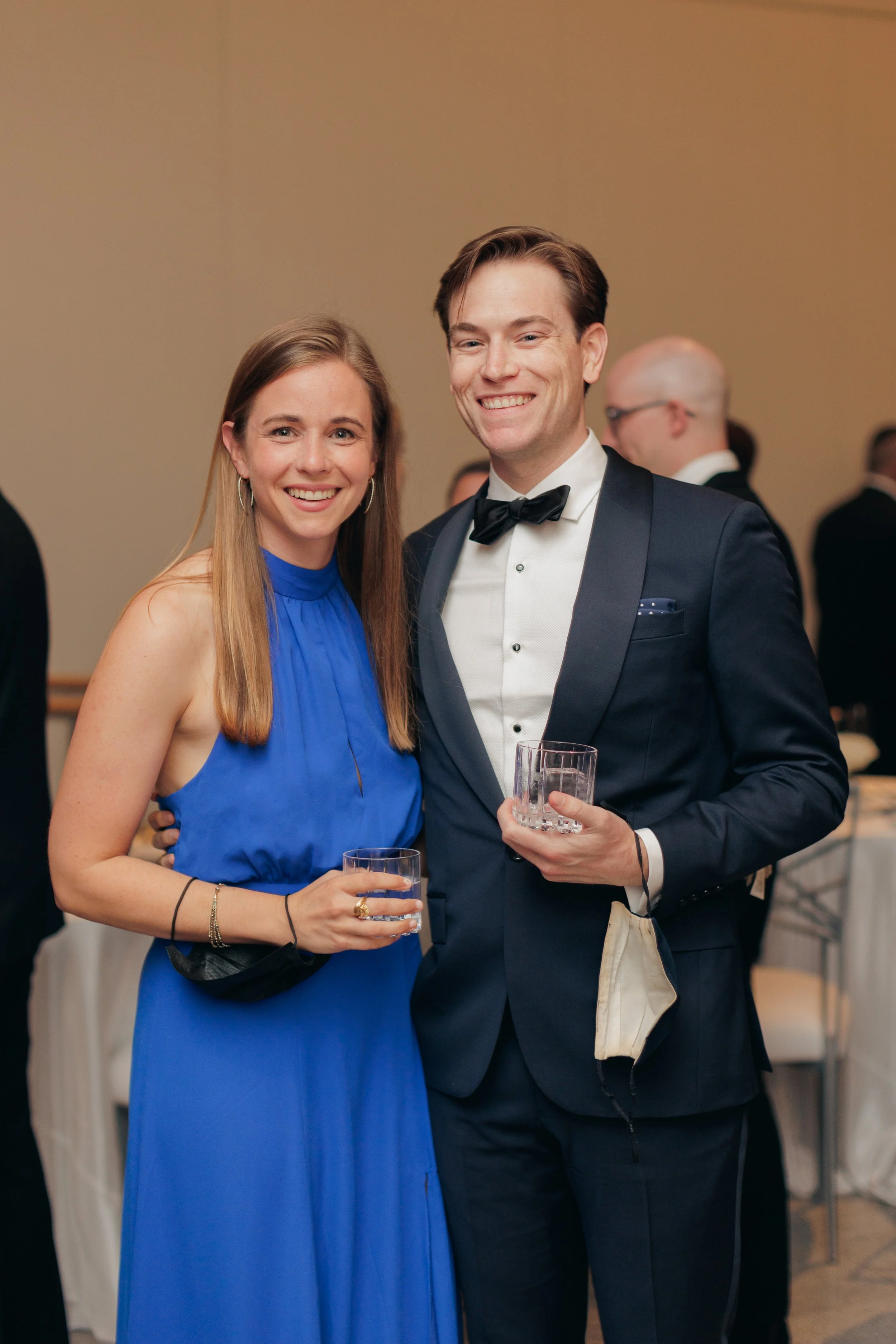 A smiling woman in a blue dress and a smiling man in formal wear, holding glasses of drink, at a formal event or gathering.