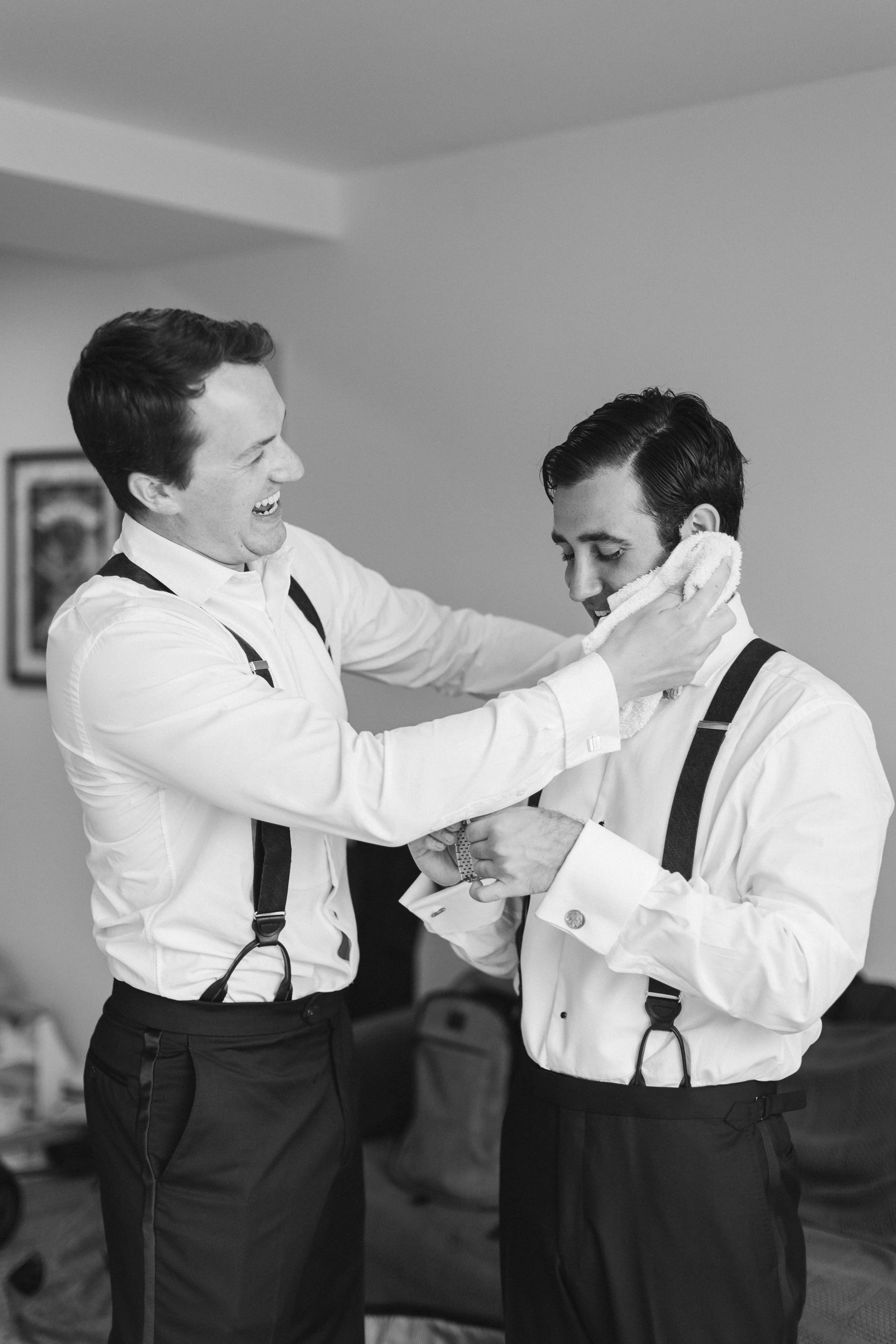 Two men dressed in white shirts, black pants, and suspenders, one helping the other adjust his cufflink, smiling with a towel draped around his neck.