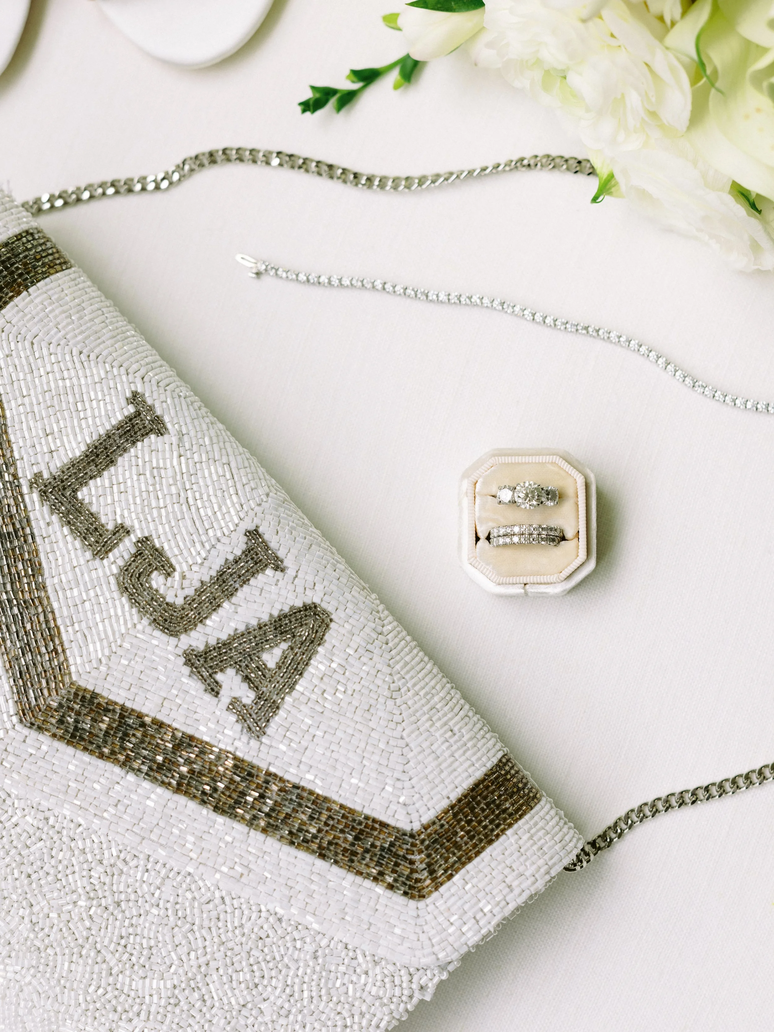 Elegant jewelry including a diamond ring, a bracelet, and a necklace displayed on a white surface with a floral arrangement and a beaded clutch.